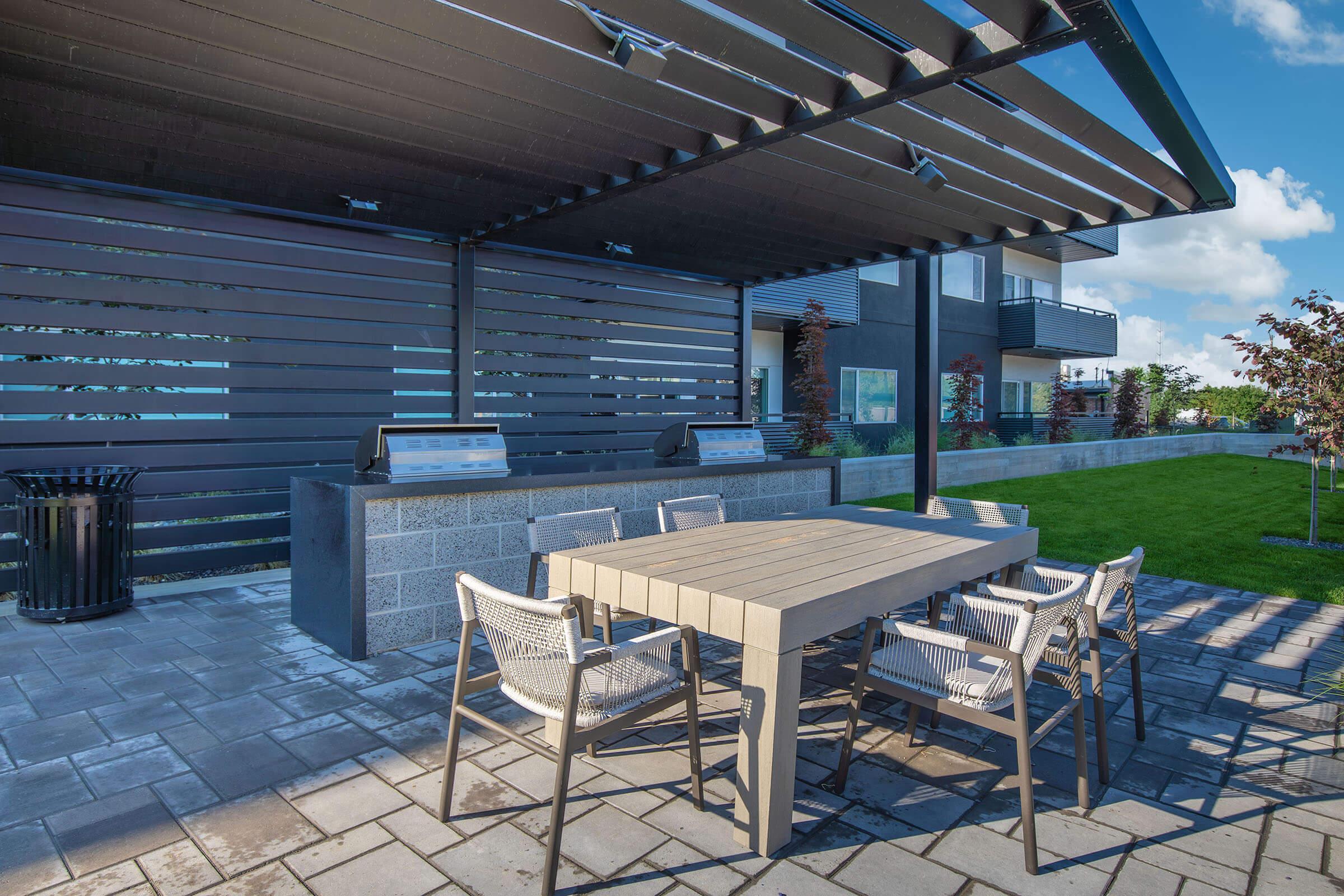 A modern outdoor patio area featuring a wooden dining table surrounded by six chairs. The space includes a sleek overhead structure with ambient lighting, and two grilling stations in the background. Lush green grass and a contemporary building are visible, creating a relaxing outdoor environment.