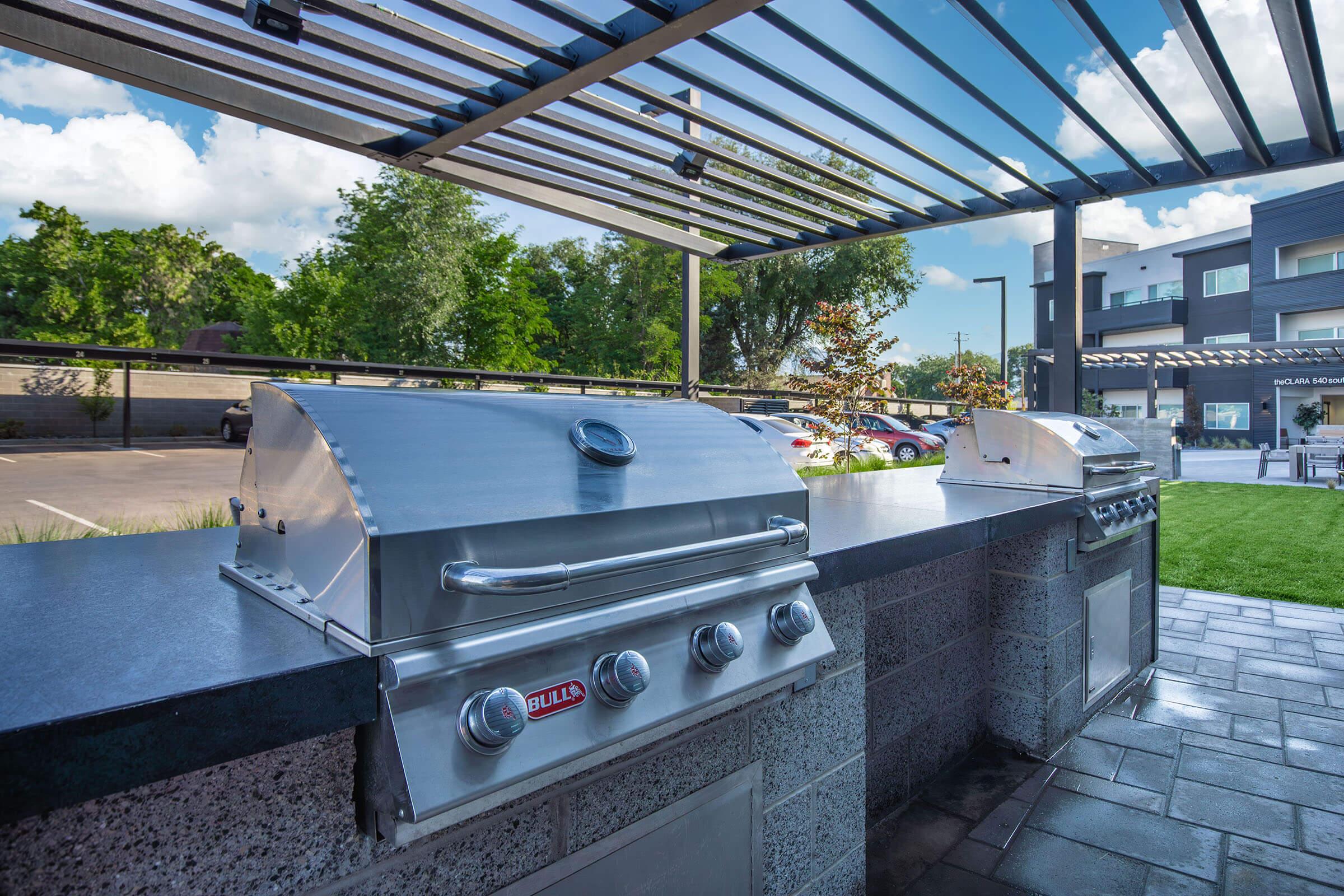 Outdoor grilling area featuring two stainless steel gas grills set on a stone countertop under a modern pergola. Lush greenery and a parking area are visible in the background, with a bright blue sky and scattered clouds above. The scene suggests a welcoming space for social gatherings or barbecues.