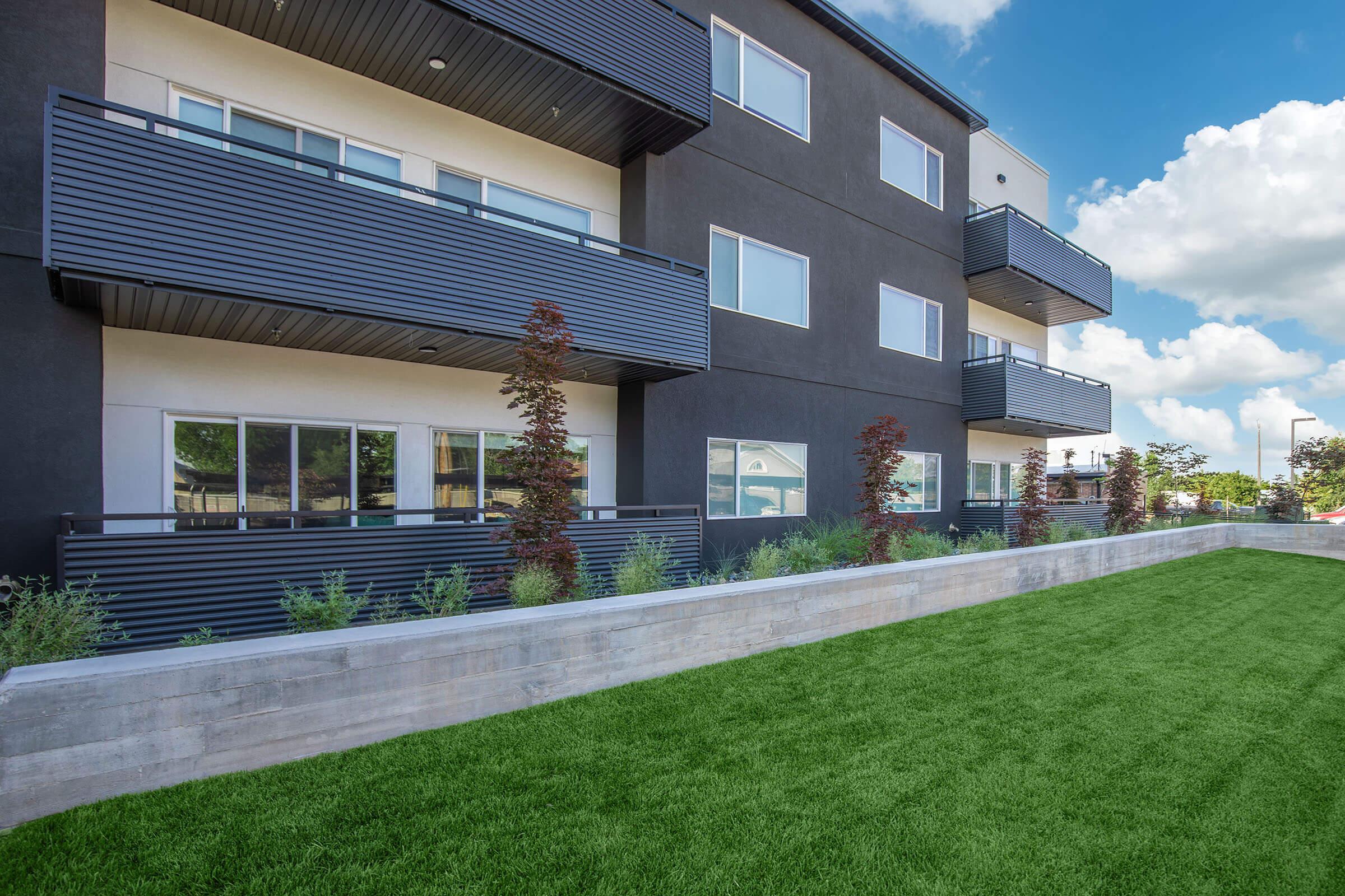 Modern apartment building with dark exterior, featuring multiple balconies. Lush green lawn in the foreground, with neatly arranged shrubs along a concrete wall. Bright blue sky with fluffy white clouds in the background, creating a welcoming atmosphere.