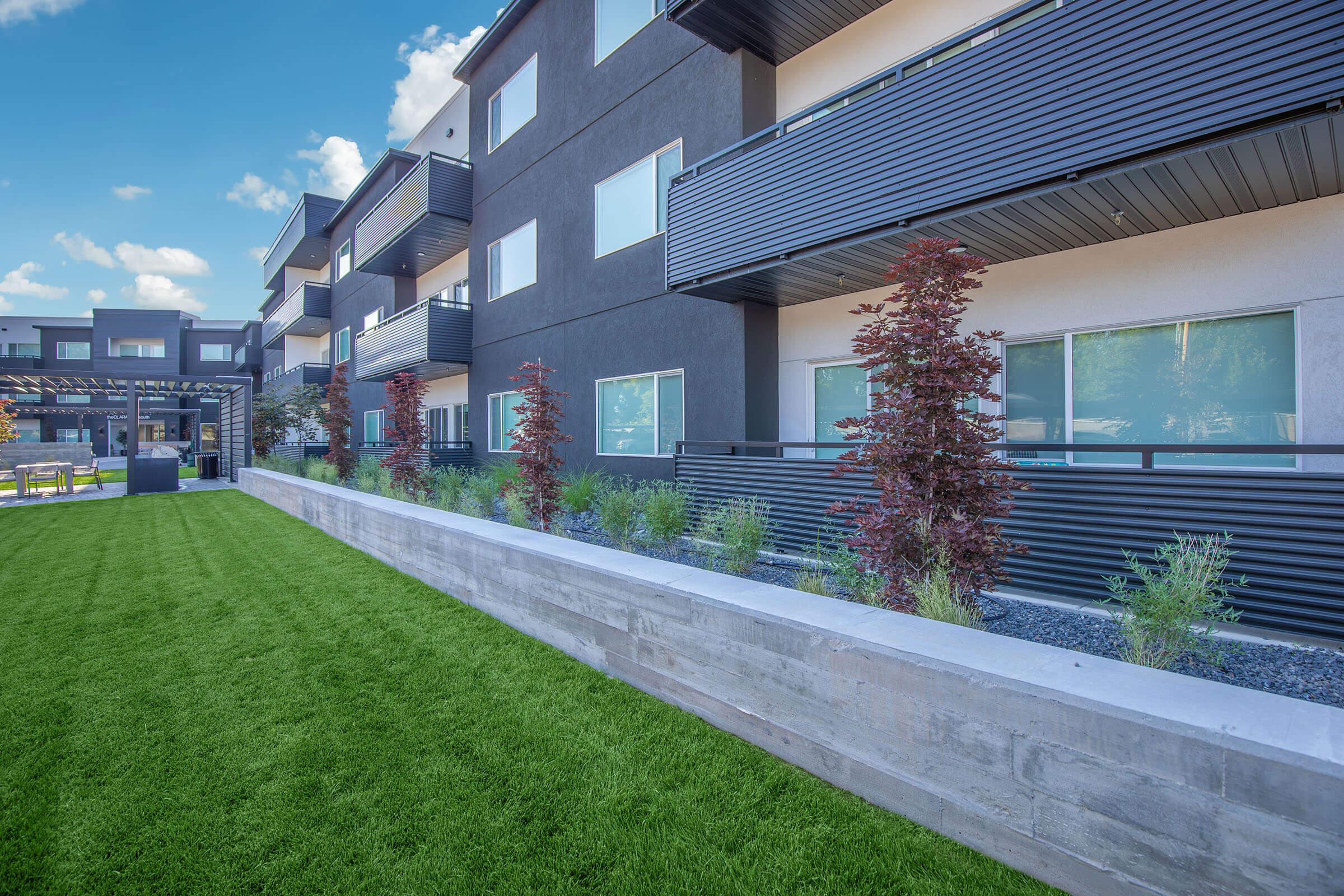 Modern apartment building exterior with multiple stories and balconies, surrounded by well-maintained green grass and ornamental plants. The design features a mix of dark and light colors, with large windows allowing natural light. Clear blue sky in the background adds to the appealing aesthetic.