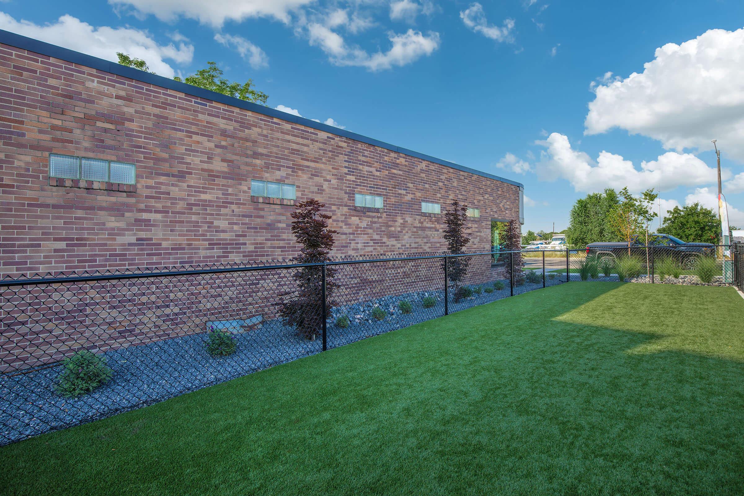 A well-maintained outdoor area featuring a brick building with several small windows. There is a green, manicured lawn bordered by a black chain-link fence. Decorative stones and shrubs are planted along the fence, complemented by a clear blue sky with scattered clouds in the background.