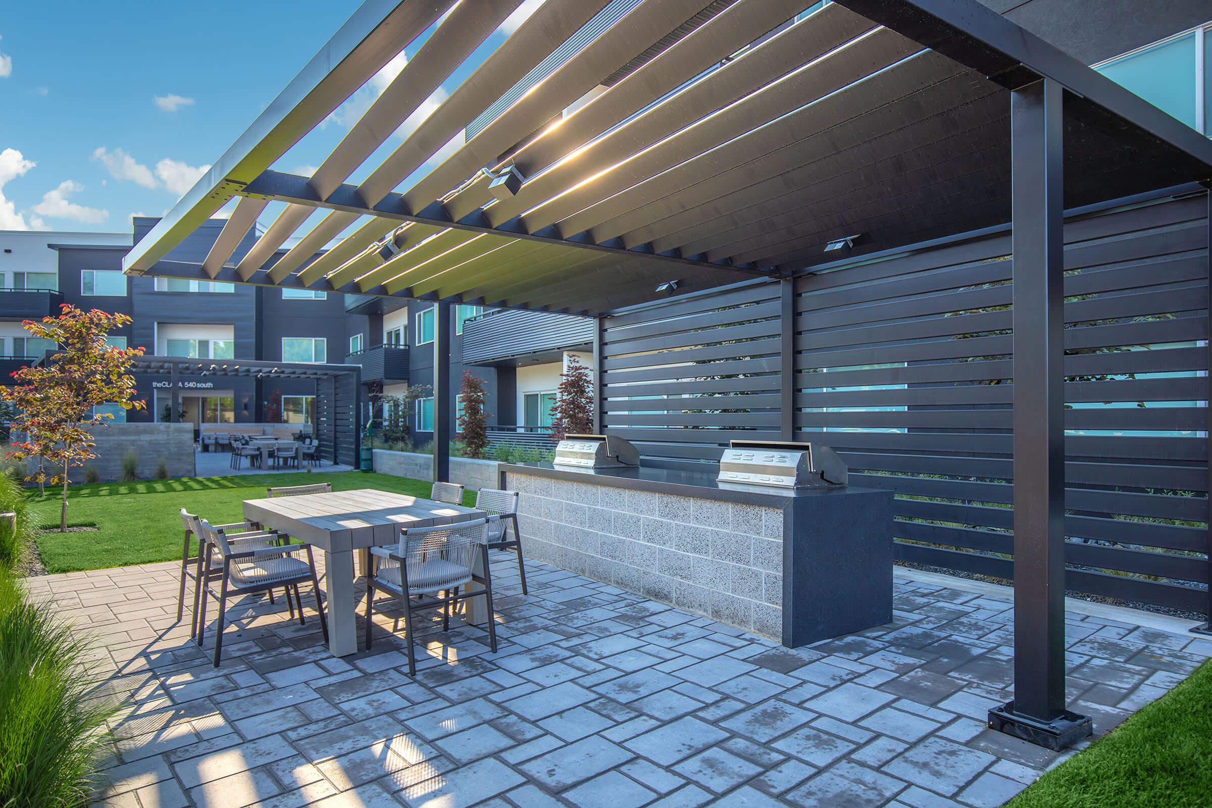 An outdoor grilling area featuring two barbecue grills under a modern pergola. The space includes a rectangular dining table with chairs, surrounded by lush green grass and landscaped elements. In the background, contemporary buildings are visible against a clear blue sky.