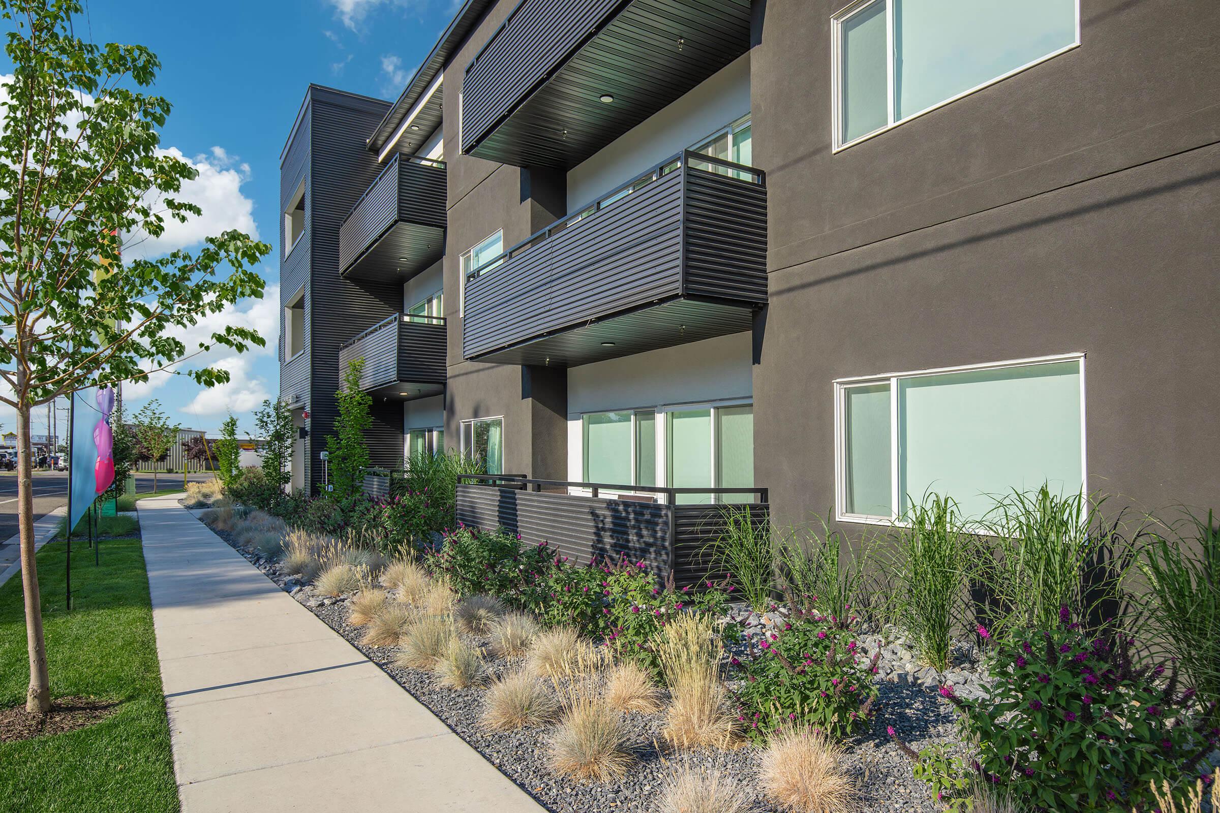 Modern apartment building with dark exterior walls and multiple balconies. The façade features large windows, surrounded by landscaped gardens with ornamental grasses and colorful flowers. A concrete sidewalk runs in front, leading through a well-maintained green space.