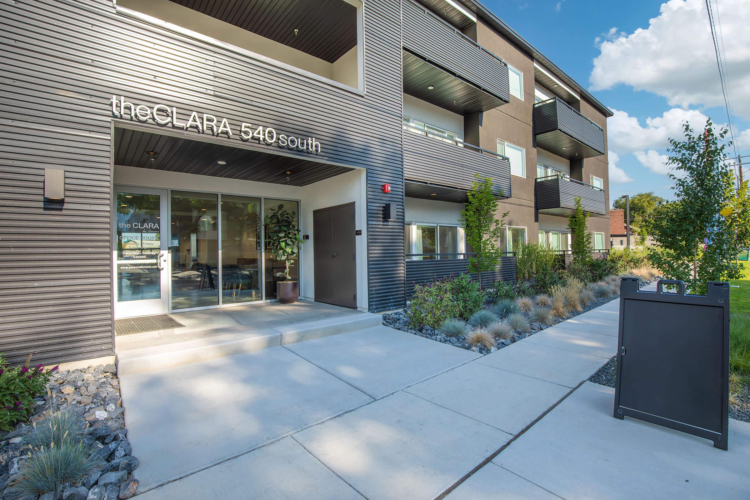 Modern apartment building entrance with large glass doors labeled "theCLARA 540 south." The exterior features sleek dark siding, green landscaping with bushes and trees, and a concrete pathway. A sign is positioned near the entrance, and the sky is blue with fluffy clouds.