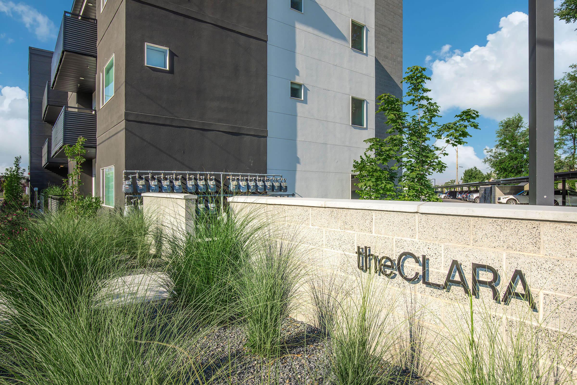 A contemporary building with a mix of dark and light gray exterior walls. In the foreground, a landscaped area with tall grasses and a stone wall featuring the name "theCLARA" in modern lettering. The background shows residential balconies and a clear blue sky with scattered clouds.