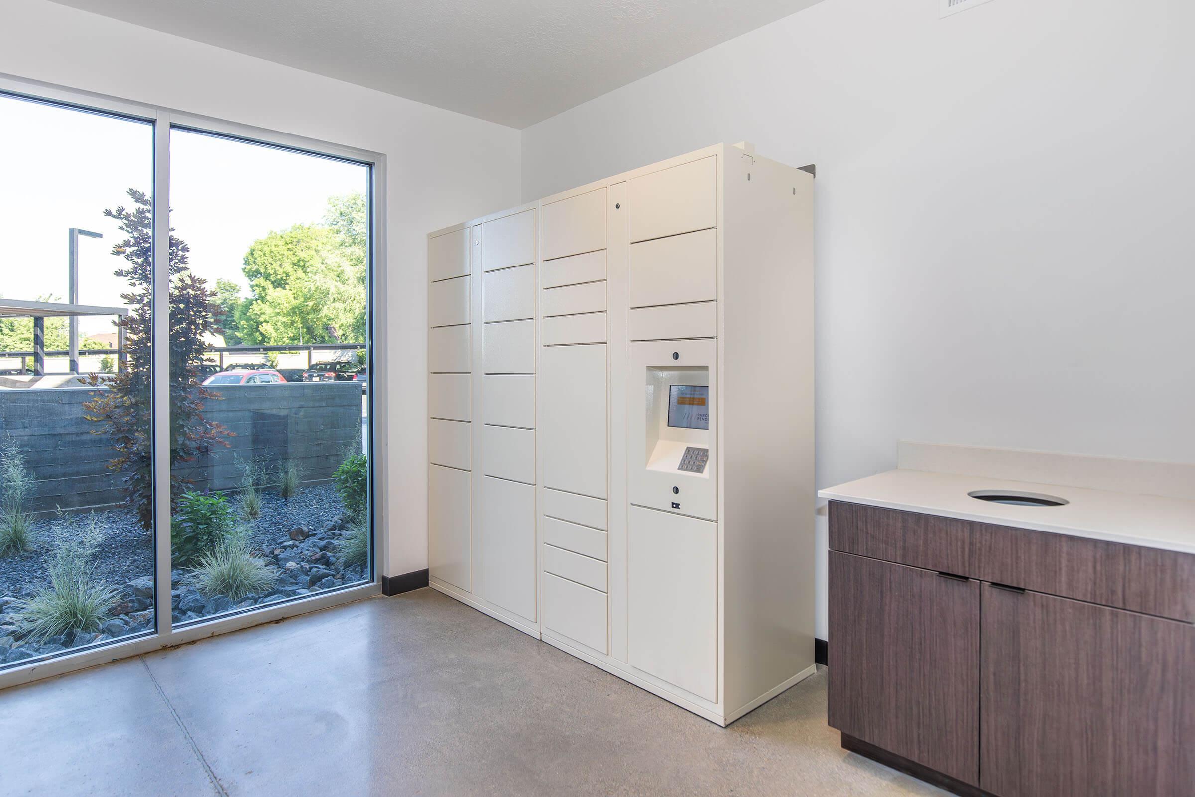 A modern room with a large window allowing natural light to enter. Inside, there is a white locker system with a touchscreen and various compartments. A countertop is visible nearby, alongside a small trash bin. Outside the window, greenery is seen, contributing to a bright and airy atmosphere.