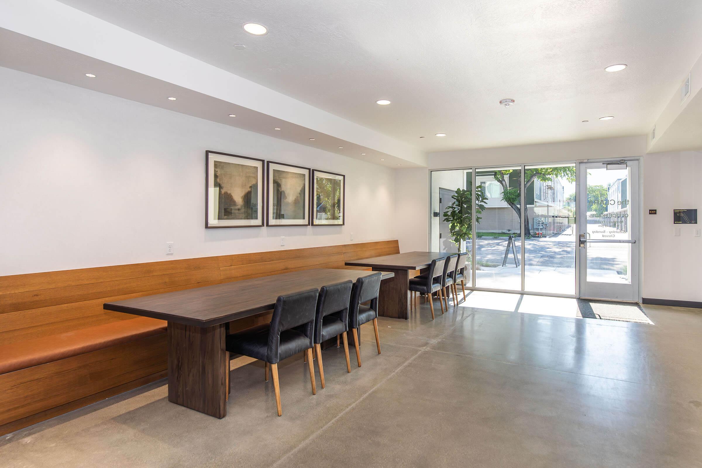 Interior of a modern room featuring a long wooden table with black chairs, a bench along one wall, and framed artwork. Large windows provide natural light and a view of the outside. The floor is concrete, and the space has a minimalist design with a neutral color palette.