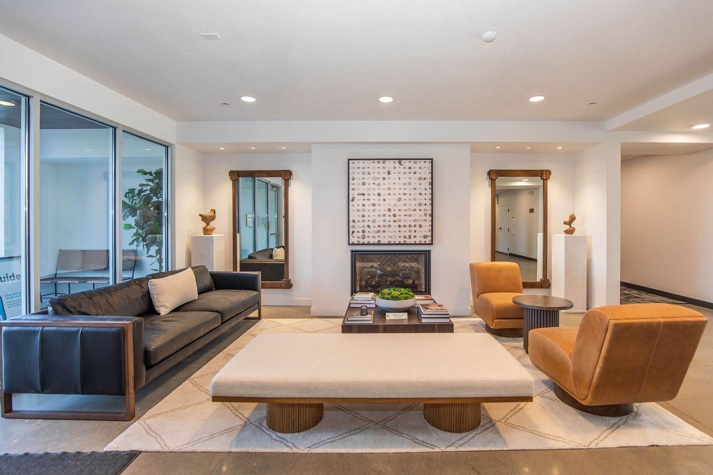 A modern, stylish lounge area featuring a black leather sofa, an orange armchair, and a light-colored coffee table. Two mirrors are mounted on the walls, reflecting the space, while a framed artwork is displayed above. Greenery adds a touch of nature, and soft lighting creates a welcoming atmosphere.