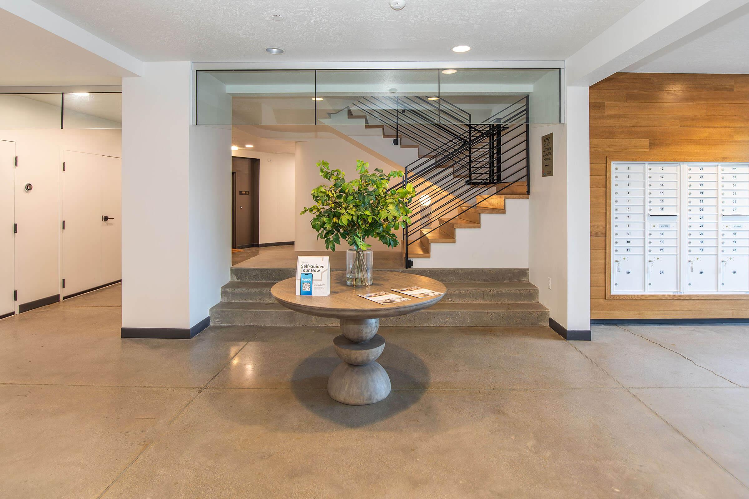 A modern lobby featuring a round table with a vase of greenery at its center. Behind the table, a staircase with a black metal railing leads to an upper level. To the right, there are several mailboxes mounted on the wall. The space has a minimalist design with a concrete floor and wooden accents.