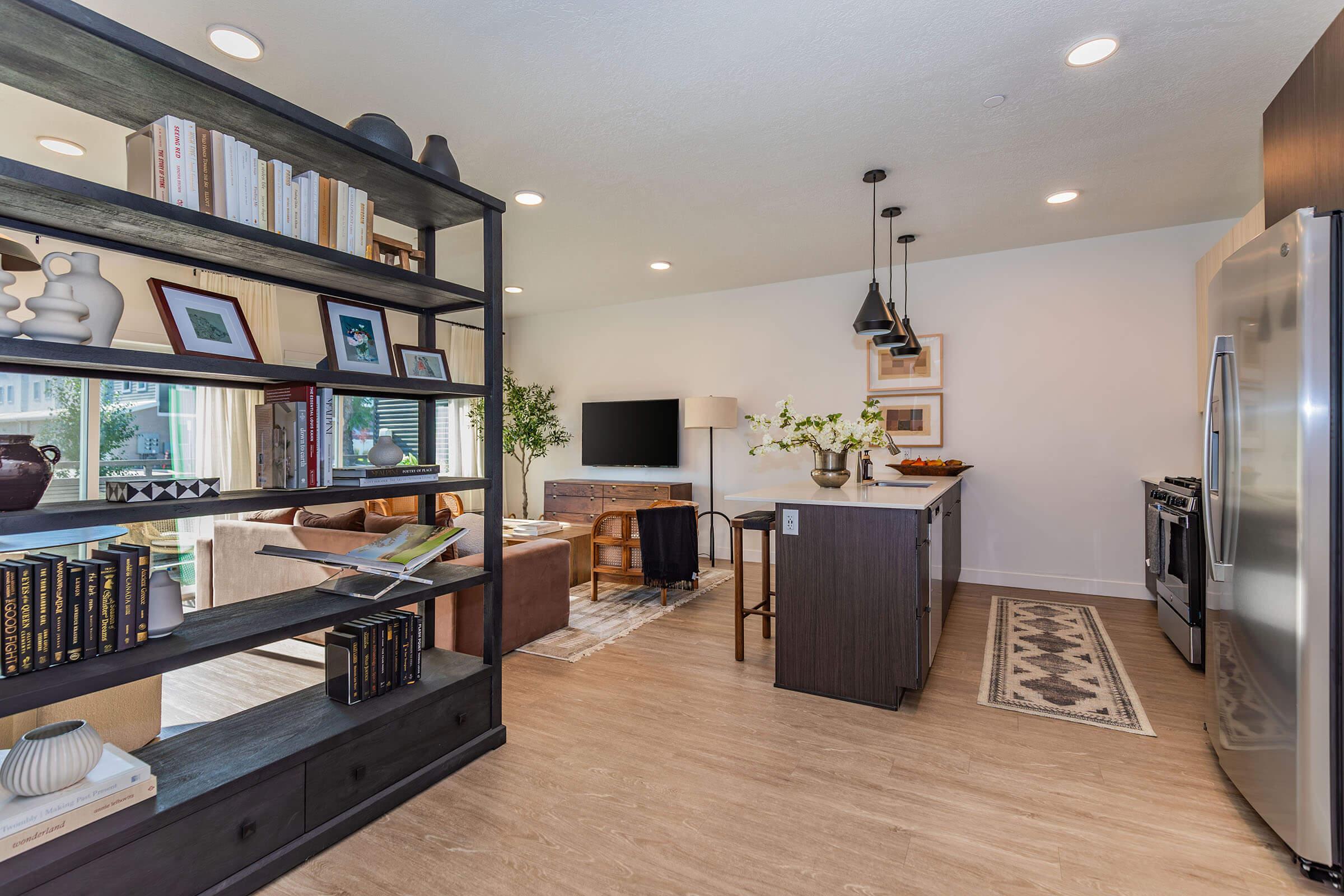 A modern, open-concept kitchen and living area featuring a dark wood shelf filled with books and decorative items. The kitchen has stainless steel appliances, a central island, and a cozy seating area with a brown sofa. Natural light filters through large windows, enhancing the contemporary decor.