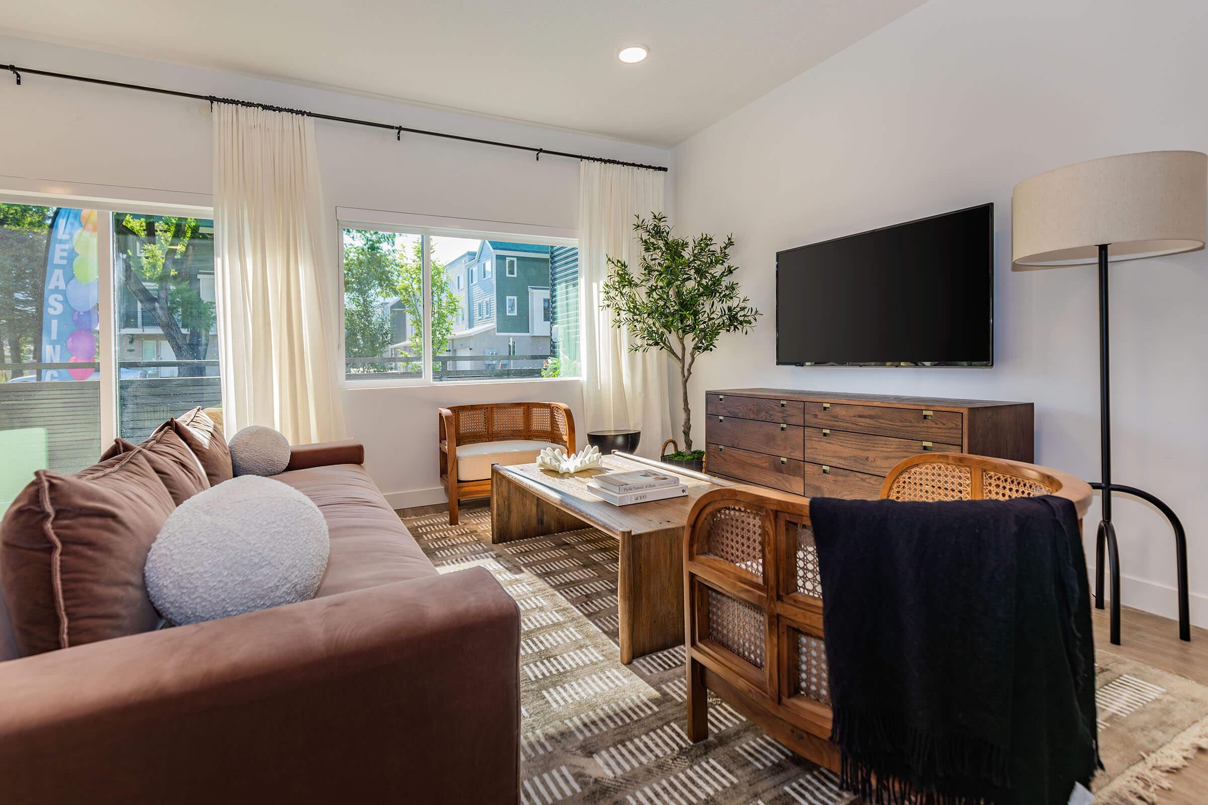 A cozy living room featuring a brown sectional sofa with decorative pillows, a wooden coffee table, and two rattan-accent chairs. A large flat-screen TV is mounted on the wall, and there are windows with sheer curtains letting in natural light. A small indoor tree adds greenery to the space.