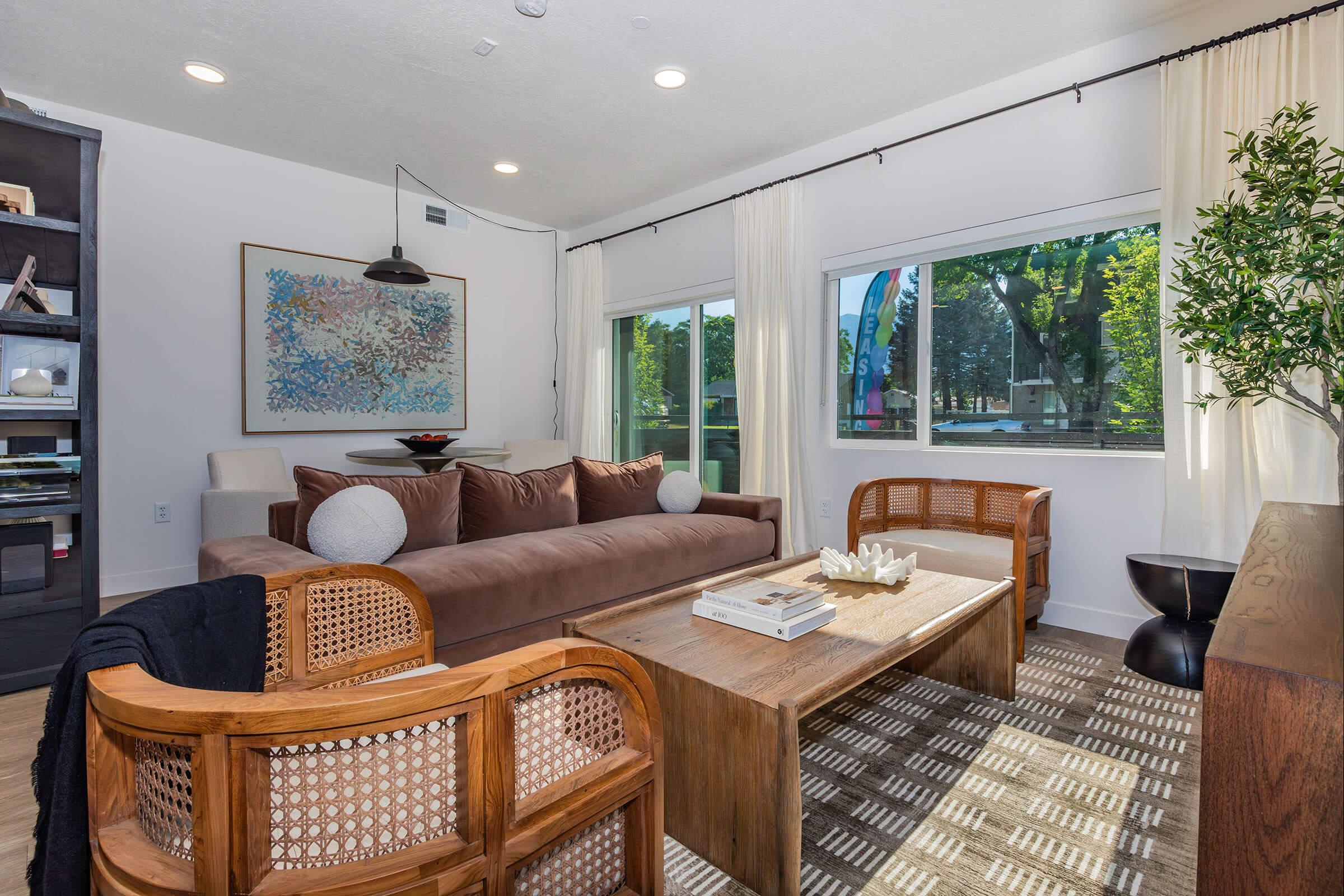 A modern living room featuring a brown sofa with decorative pillows, two wooden chairs, a wooden coffee table, and a large artwork on the wall. Natural light enters through large windows adorned with sheer curtains, highlighting the cozy, stylish atmosphere.