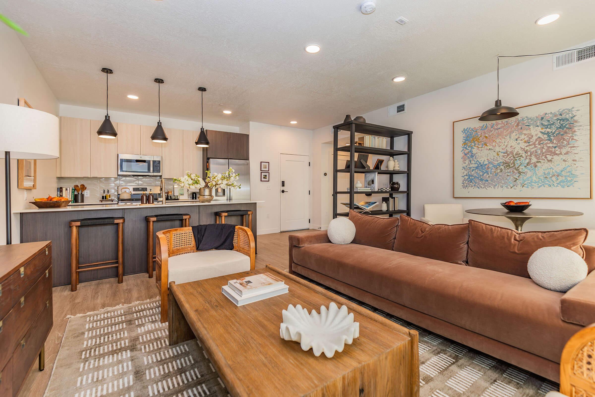 A modern living room featuring a brown sofa, a wooden coffee table, and stylish decor. The open kitchen in the background includes stainless steel appliances. Pendant lights hang above the kitchen island, complemented by a decorative bookshelf. Soft textures and neutral colors create a cozy ambiance.