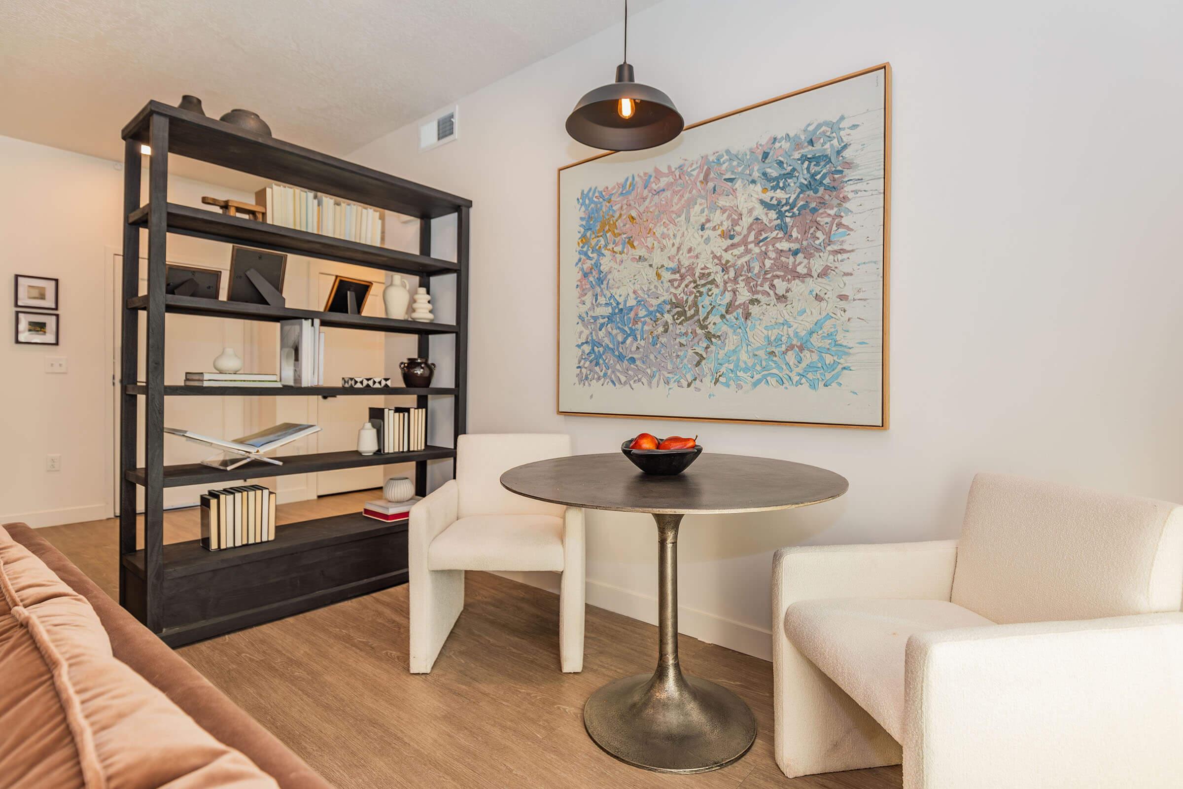 A cozy interior scene featuring a modern bookshelf filled with books and decorative items, a round table with a bowl of fruit, and two white upholstered chairs. A colorful abstract painting hangs on the wall, complemented by stylish lighting, creating a warm and inviting atmosphere.