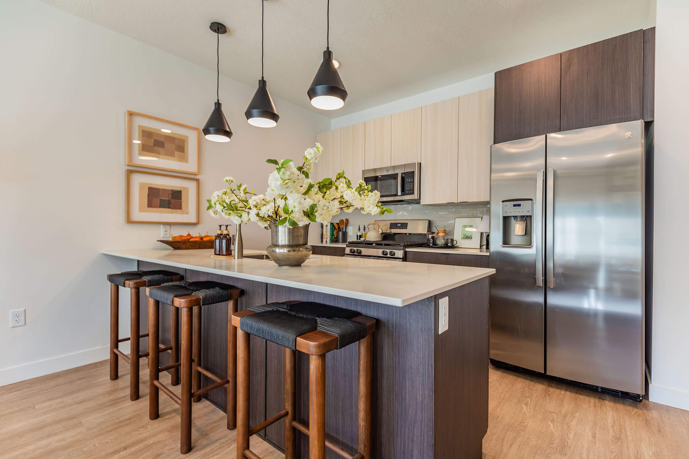 Modern kitchen featuring a large island with four wooden stools, stainless steel appliances, and decorative vases with flowers. The cabinetry combines light wood and dark finishes. Pendant lights hang above the island, creating a stylish and inviting atmosphere. Wall art complements the contemporary design.