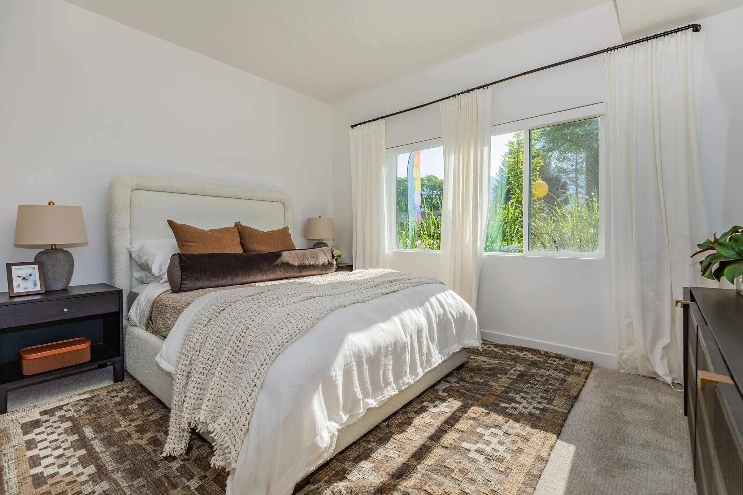 A bright and airy bedroom featuring a plush bed with decorative pillows, a woven blanket, and a textured rug. Large windows allow natural light to fill the room, framed by sheer white curtains. A bedside table with a lamp and a small decorative item, along with a potted plant, add warmth to the space.