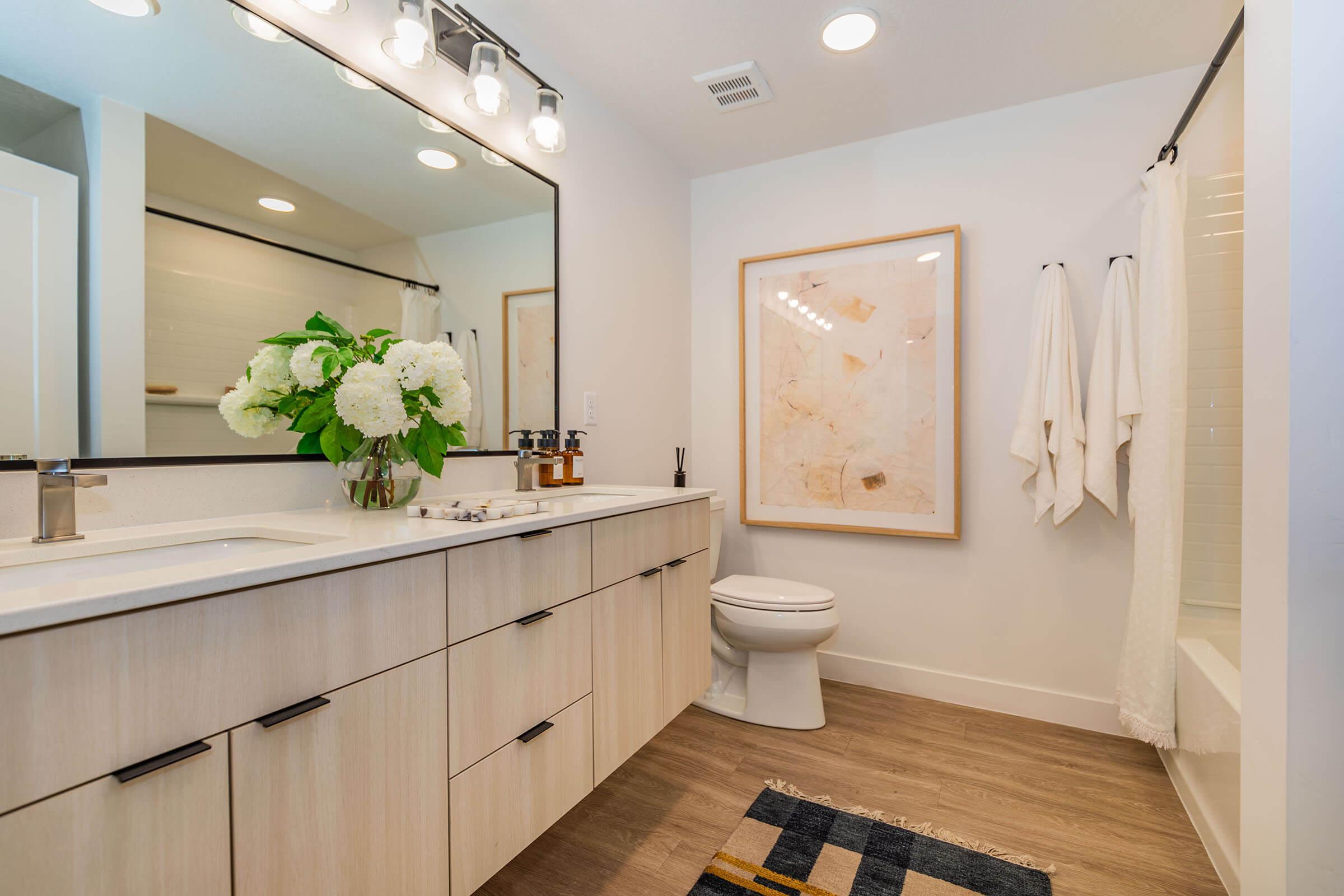 Modern bathroom featuring a double vanity with light wood cabinetry, a large mirror, and stylish fixtures. A bouquet of white flowers sits on the countertop. The space includes cream-colored towels hanging on hooks, a framed abstract art piece on the wall, and a bathtub/shower combination with a light tile backdrop.