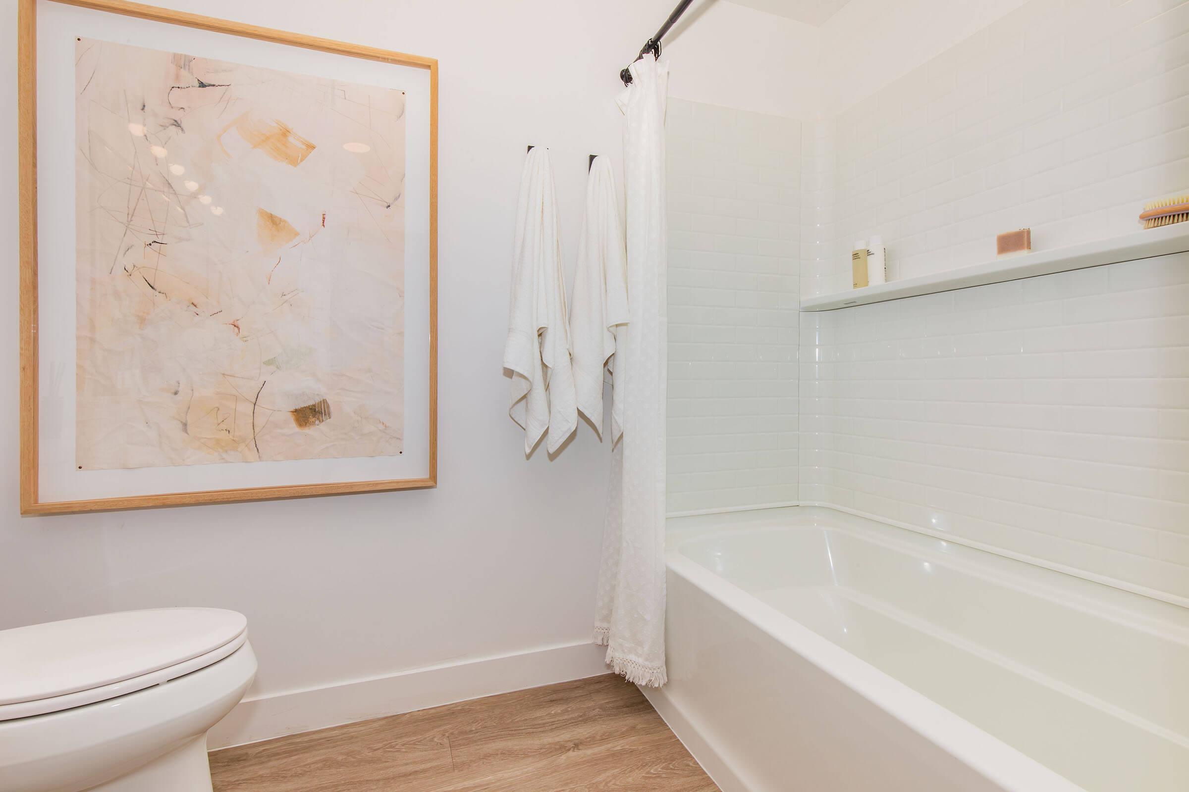 A modern, bright bathroom featuring a white bathtub, a framed piece of abstract art on the wall, and two white towels hanging on a rack. The floor is styled with light-colored wood, adding warmth to the space. Natural light brightens the area, creating a clean and inviting atmosphere.