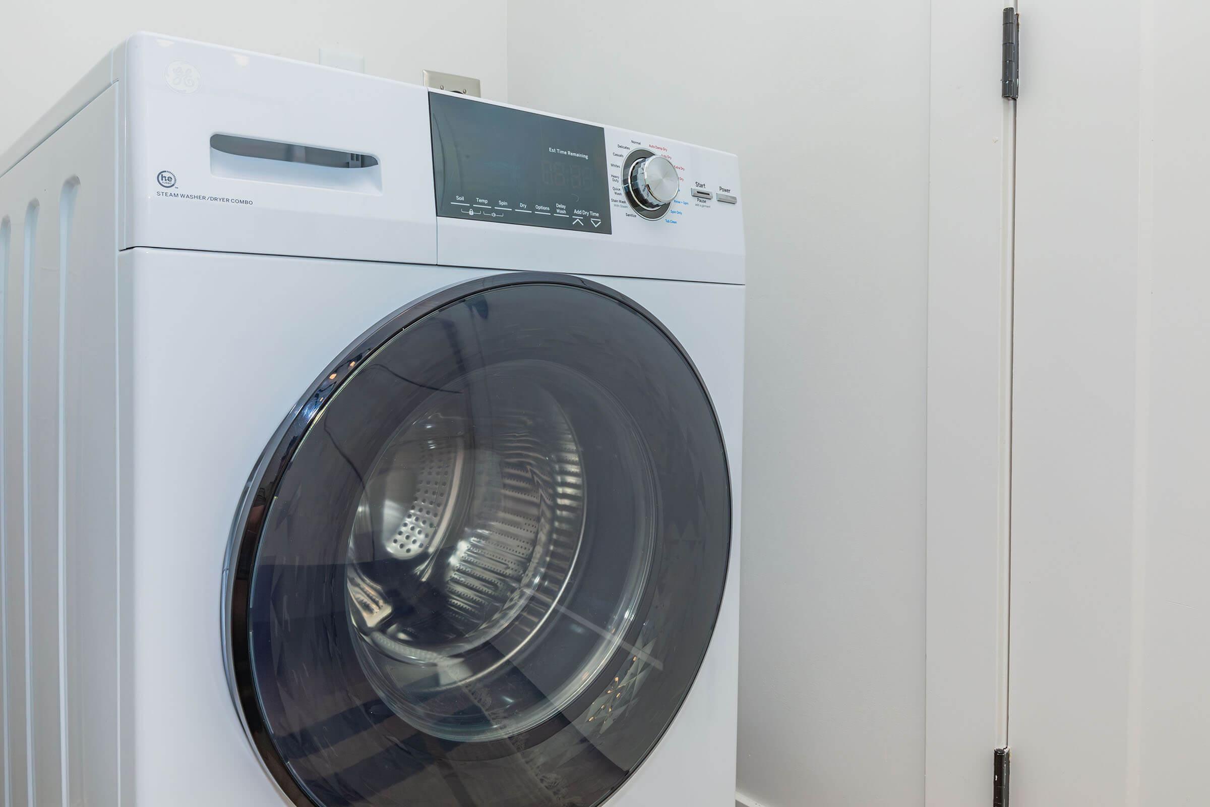 A modern white front-loading washing machine is positioned against a light-colored wall. The machine features a large, tinted glass door, a control panel with various settings, and a sleek, minimalist design. A closed door is visible in the background, contributing to a clean and organized laundry space.