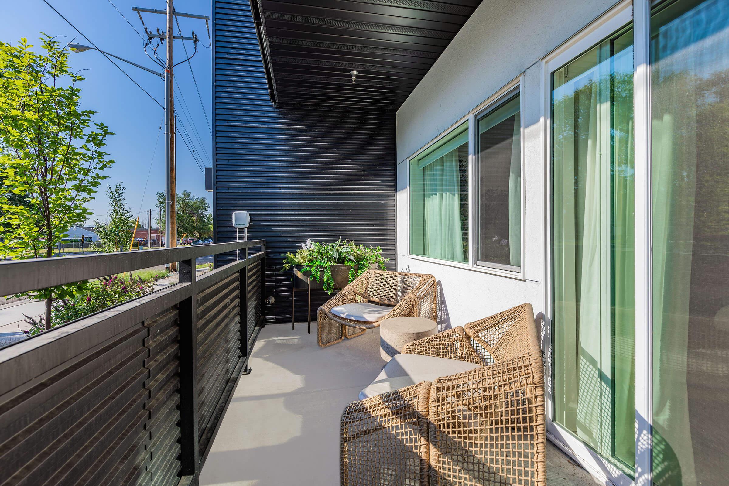 A modern balcony featuring two wicker chairs and a small table, surrounded by green plants. The balcony has a sleek black railing and large glass windows, allowing natural light to enter the space. Power lines and trees are visible in the background, suggesting an urban setting.