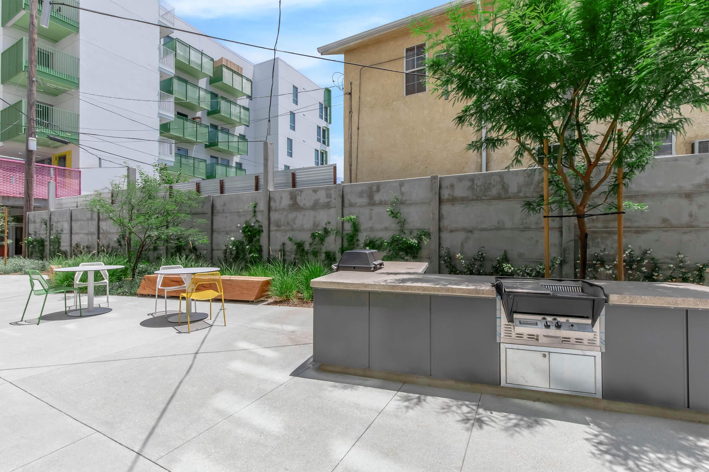 Outdoor communal space featuring a grill area with a barbecue, surrounded by plants and trees. There are modern tables and chairs in vibrant colors, with a concrete wall in the background and apartment buildings visible beyond. A bright and inviting environment for social gatherings.