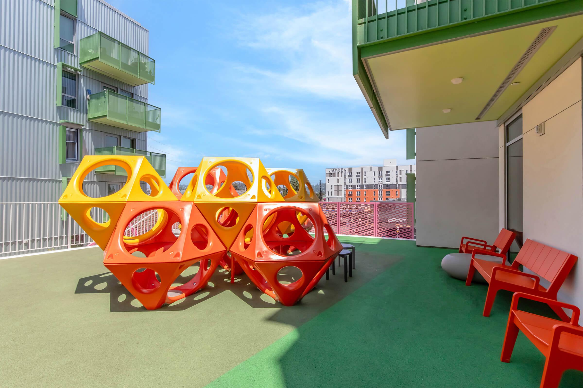 Colorful geometric play structure made of orange and yellow, situated on a rooftop terrace. Red outdoor chairs are placed nearby, with a modern building and blue sky in the background. The space appears designed for children's play and relaxation.
