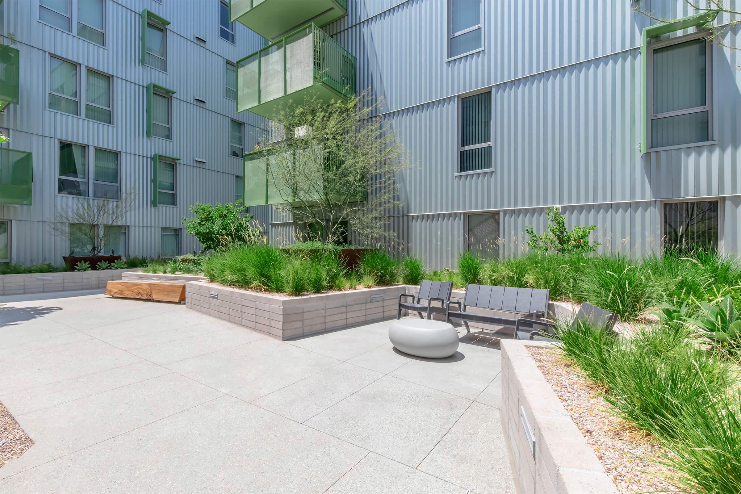 A modern outdoor courtyard featuring low seating, a circular bench, and lush greenery. The surroundings include stylish building facades with metal siding and large windows. Natural light fills the space, creating a serene atmosphere perfect for relaxation.