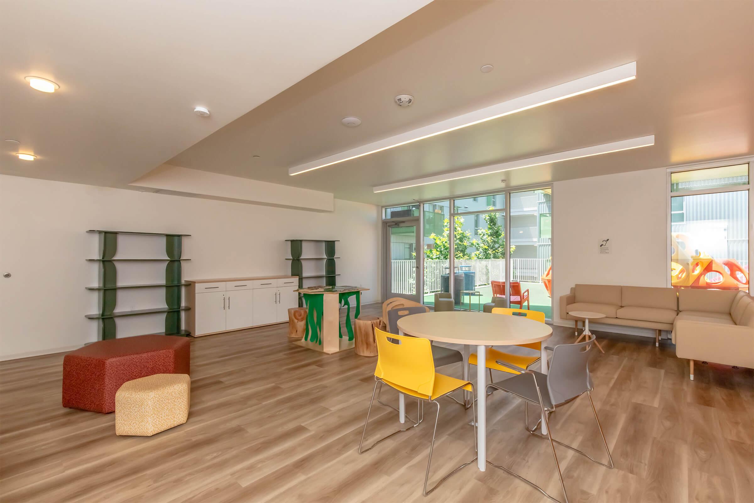A spacious, modern community room featuring a round table with colorful chairs, a cozy couch, and various shelves. The room is bright with natural light coming through large windows, offering a casual, inviting atmosphere for gatherings or activities.