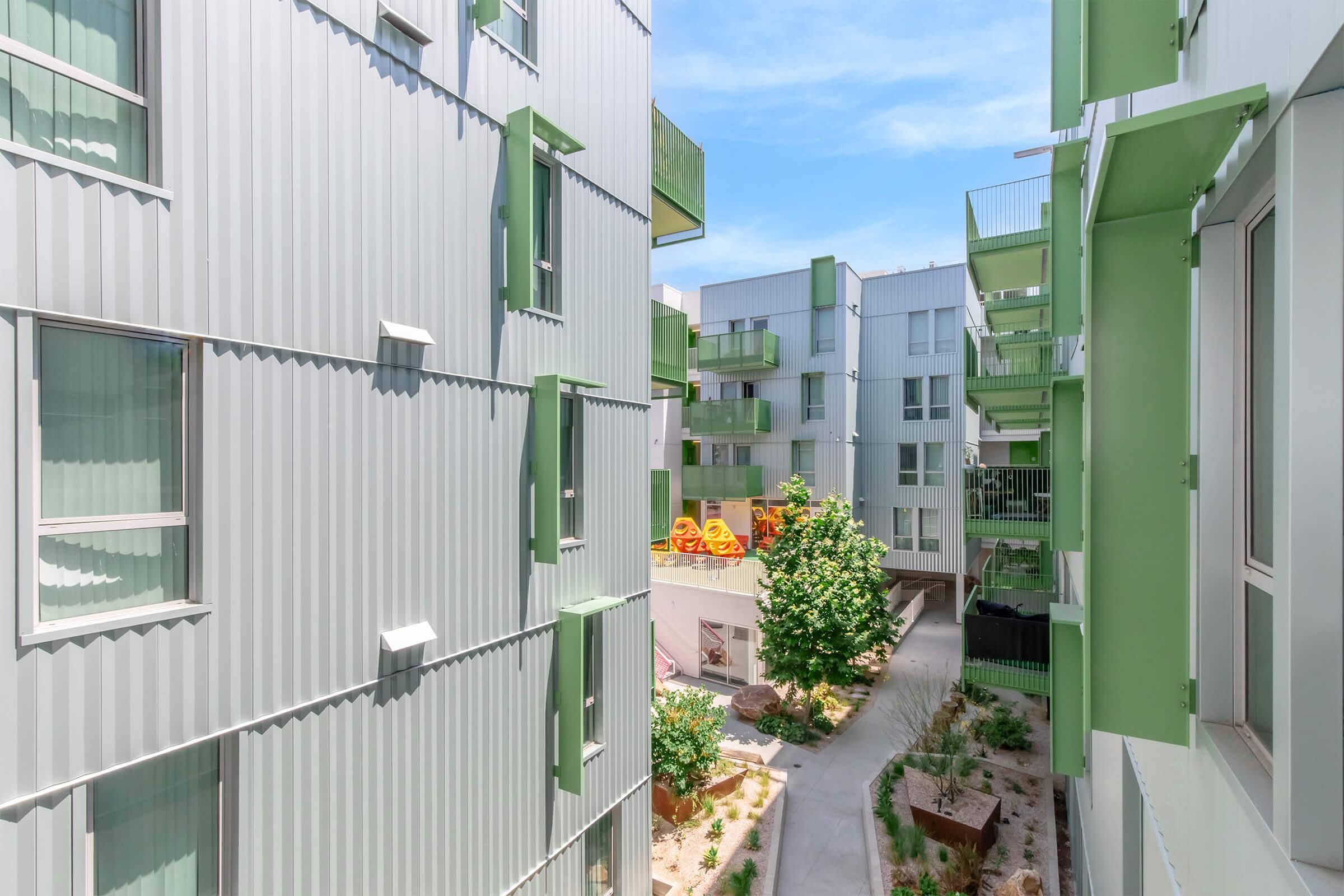 View of a modern residential complex with corrugated metal exteriors. The buildings feature green accents and balconies, surrounding a landscaped courtyard with a tree and planters. Bright blue sky is visible above, enhancing the contemporary architectural design.