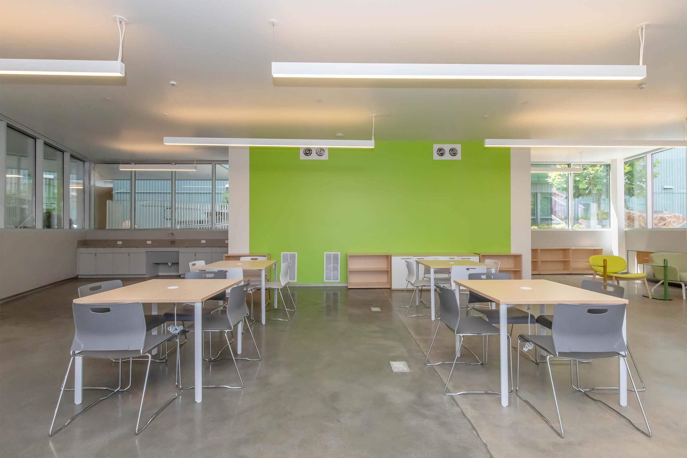 A modern classroom with light gray concrete flooring, featuring multiple wooden tables and chairs arranged neatly. The walls are mostly white with one bright green accent wall. Large windows allow natural light to fill the space, and there are air vents and shelves visible. The overall design is minimalistic and bright.