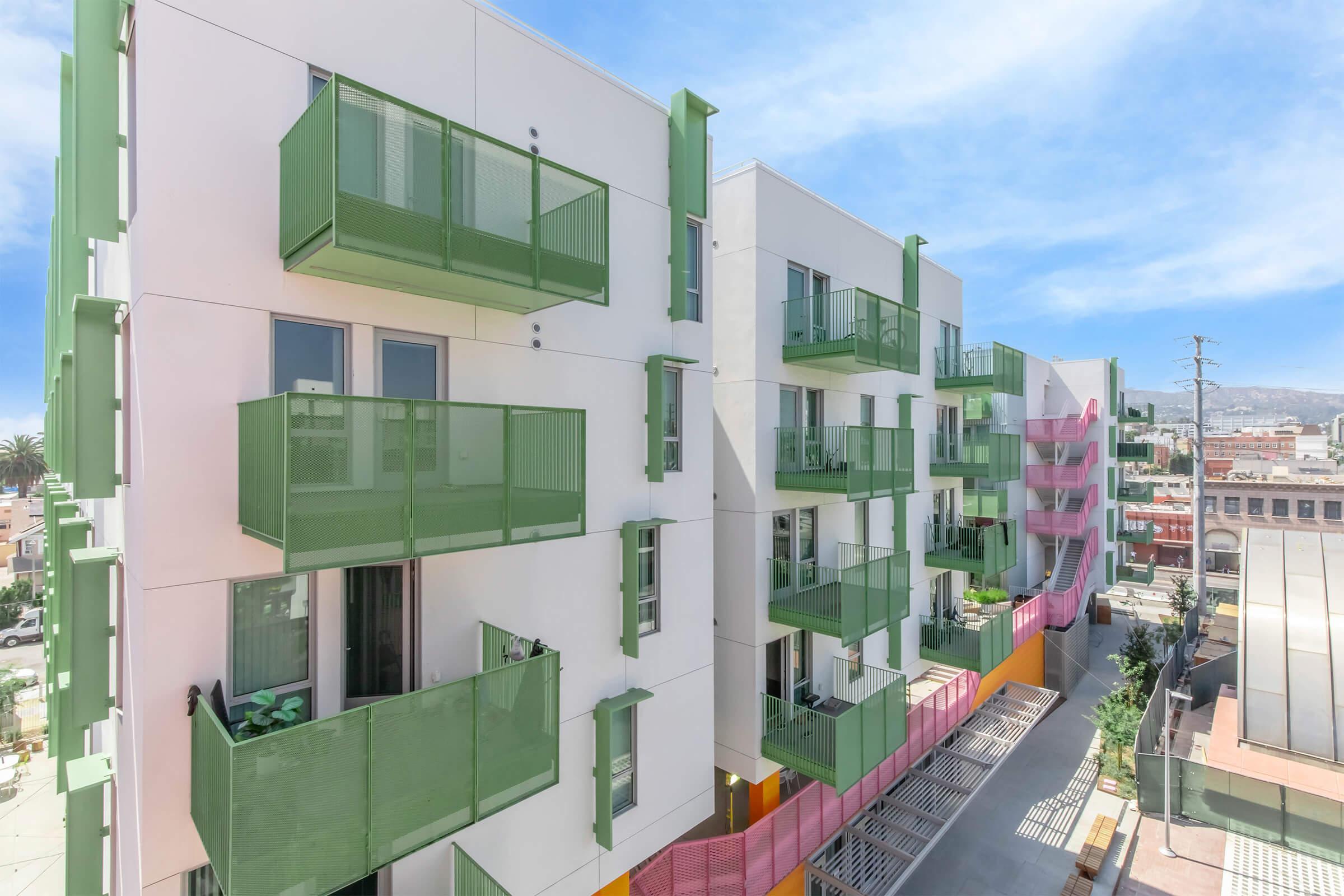 A modern apartment building featuring multiple stories with green balconies and colorful accents. The architecture is contemporary, showcasing a mix of white walls and vibrant details, set against a bright blue sky. Urban surroundings with nearby buildings and a clear view of the street below can be seen.