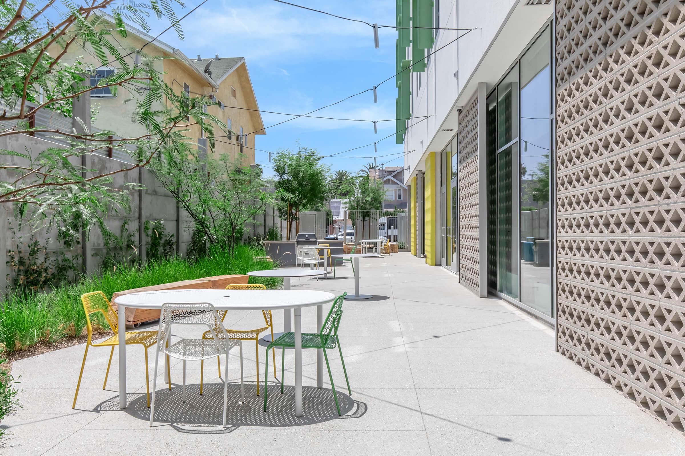 A well-lit outdoor area featuring tables and chairs in various colors. Green plants and trees border the space, with modern buildings in the background. String lights hang overhead, creating a vibrant and inviting atmosphere suitable for gatherings or relaxation.