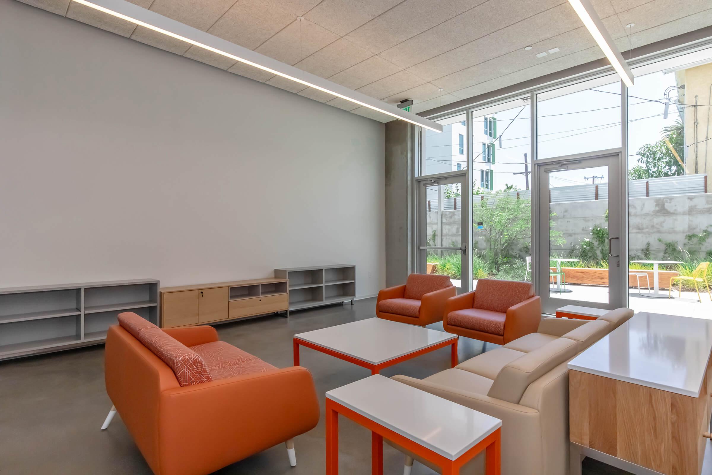 A modern indoor lounge area featuring two orange chairs, a light-colored sofa, and several coffee tables. A large, empty wall and floor-to-ceiling windows provide natural light, with outdoor greenery visible beyond the glass. Minimalistic furniture and a neutral color palette create a contemporary atmosphere.