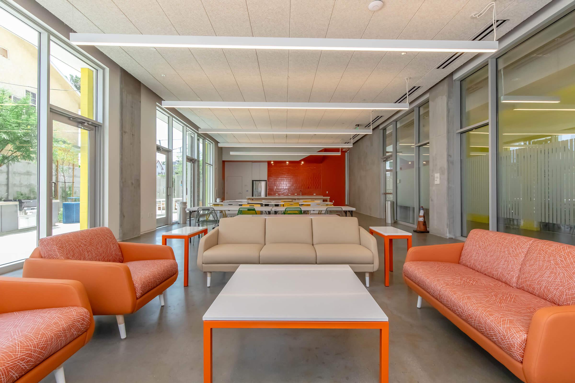 Modern interior of a spacious lounge area featuring orange sofas, a white coffee table, and large windows allowing natural light. The walls are adorned with a bright orange accent, and there are communal tables in the background, creating a vibrant and inviting atmosphere.