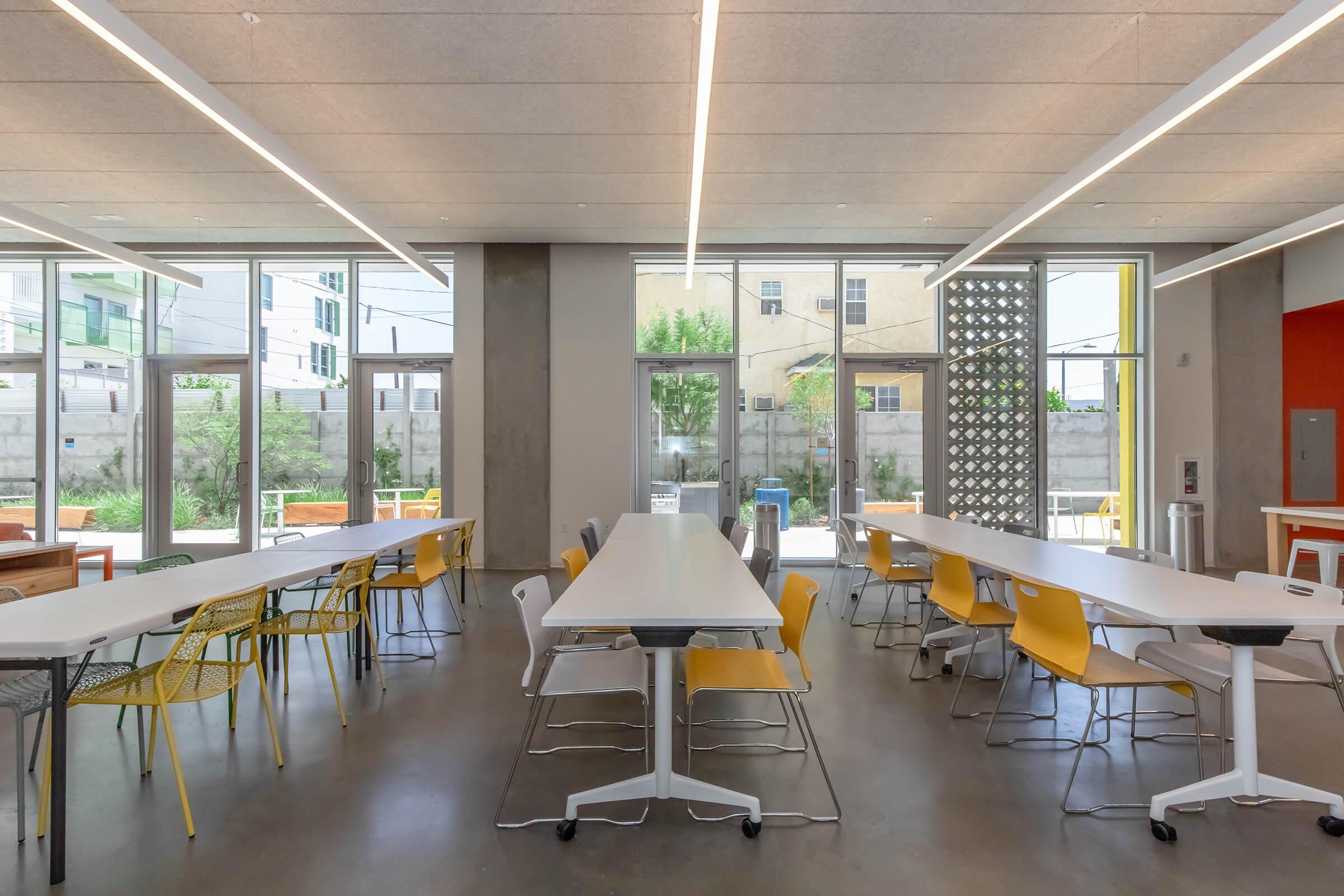 Modern workspace featuring several long white tables surrounded by yellow chairs. Large windows allow natural light to illuminate the space, with plants visible outside. The decor is sleek and minimalistic, and there are light fixtures hanging from the ceiling, creating a welcoming environment.