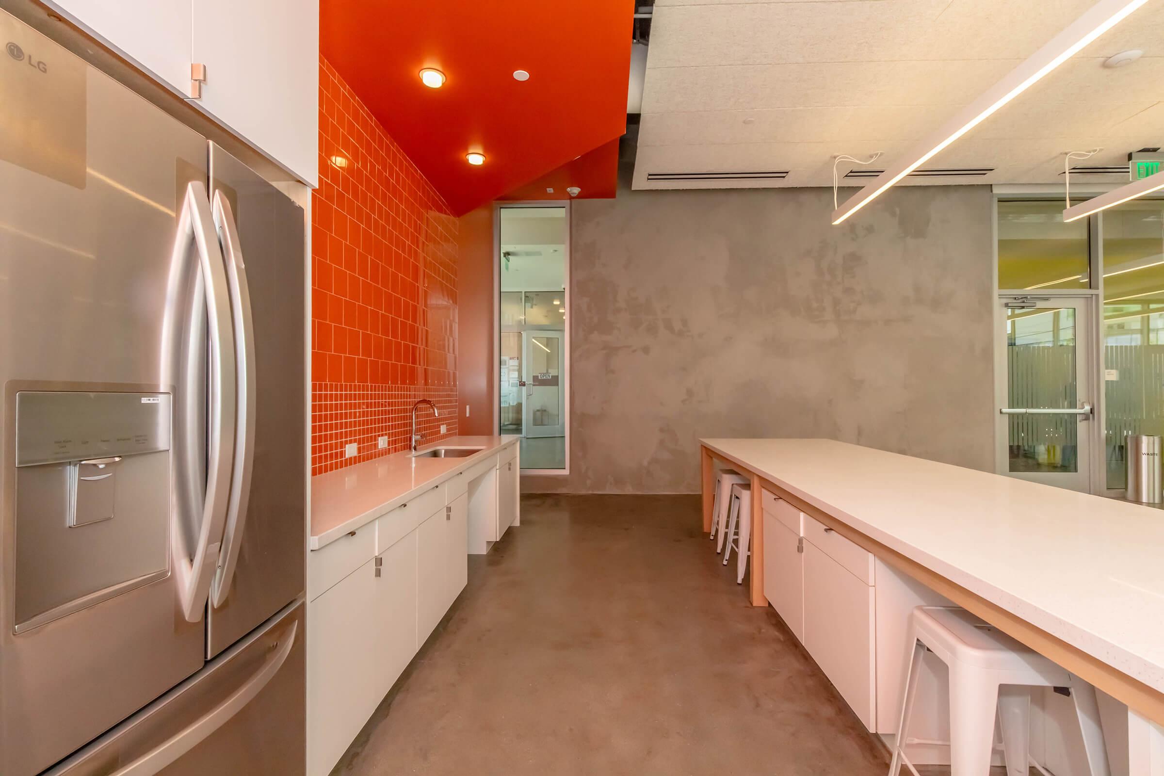 Modern kitchen space featuring stainless steel refrigerator, white countertops, and cabinets. The wall is adorned with bright orange tiles, while the floor has a concrete finish. Minimalist design with bar stools and natural lighting from adjacent glass-walled area.