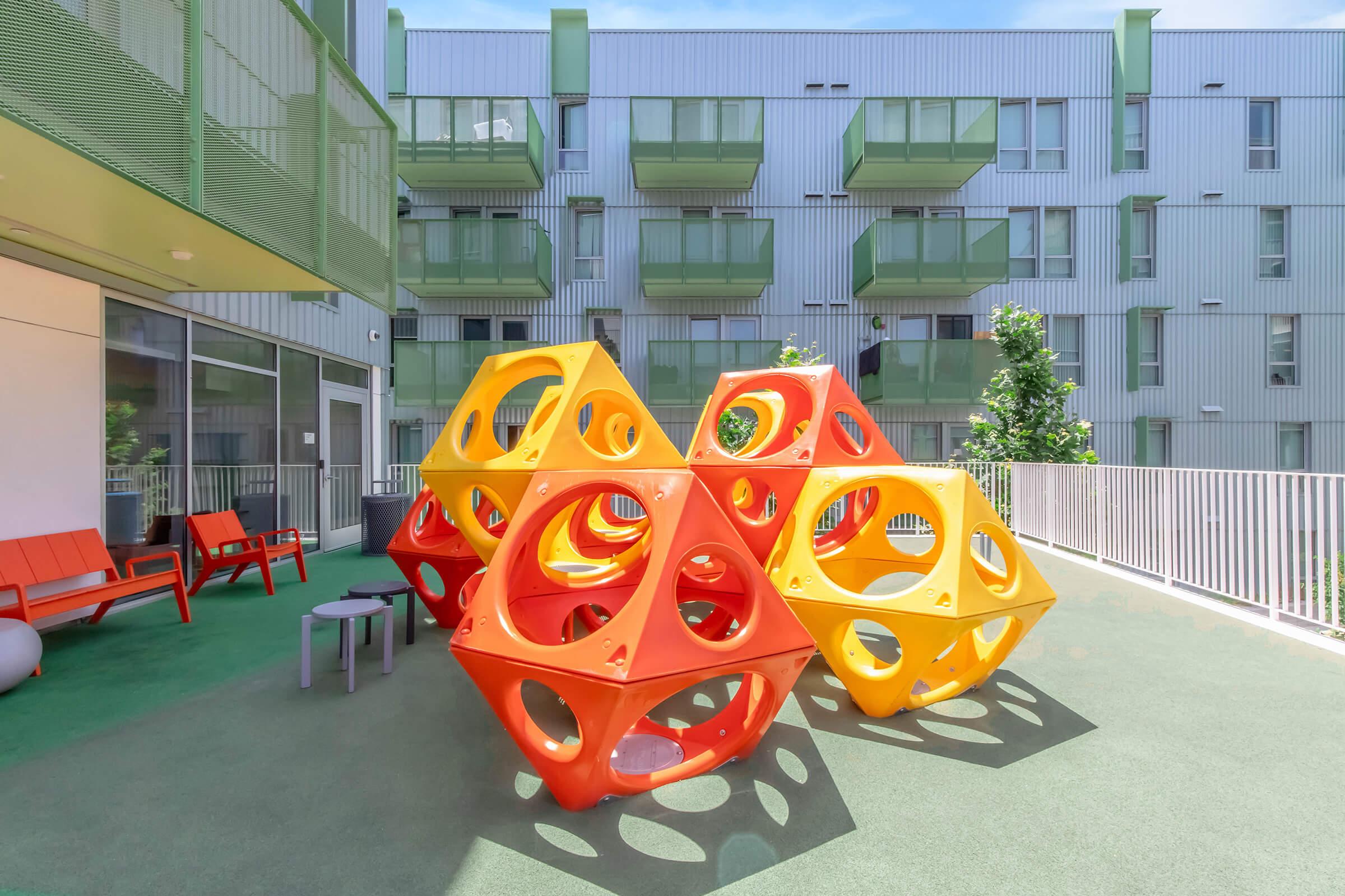Colorful outdoor playground structure made of interconnected orange and yellow geometric shapes, set in a bright, sunny courtyard with modern apartment buildings in the background. Green foliage and seating areas are visible, creating a lively and inviting play space for children.