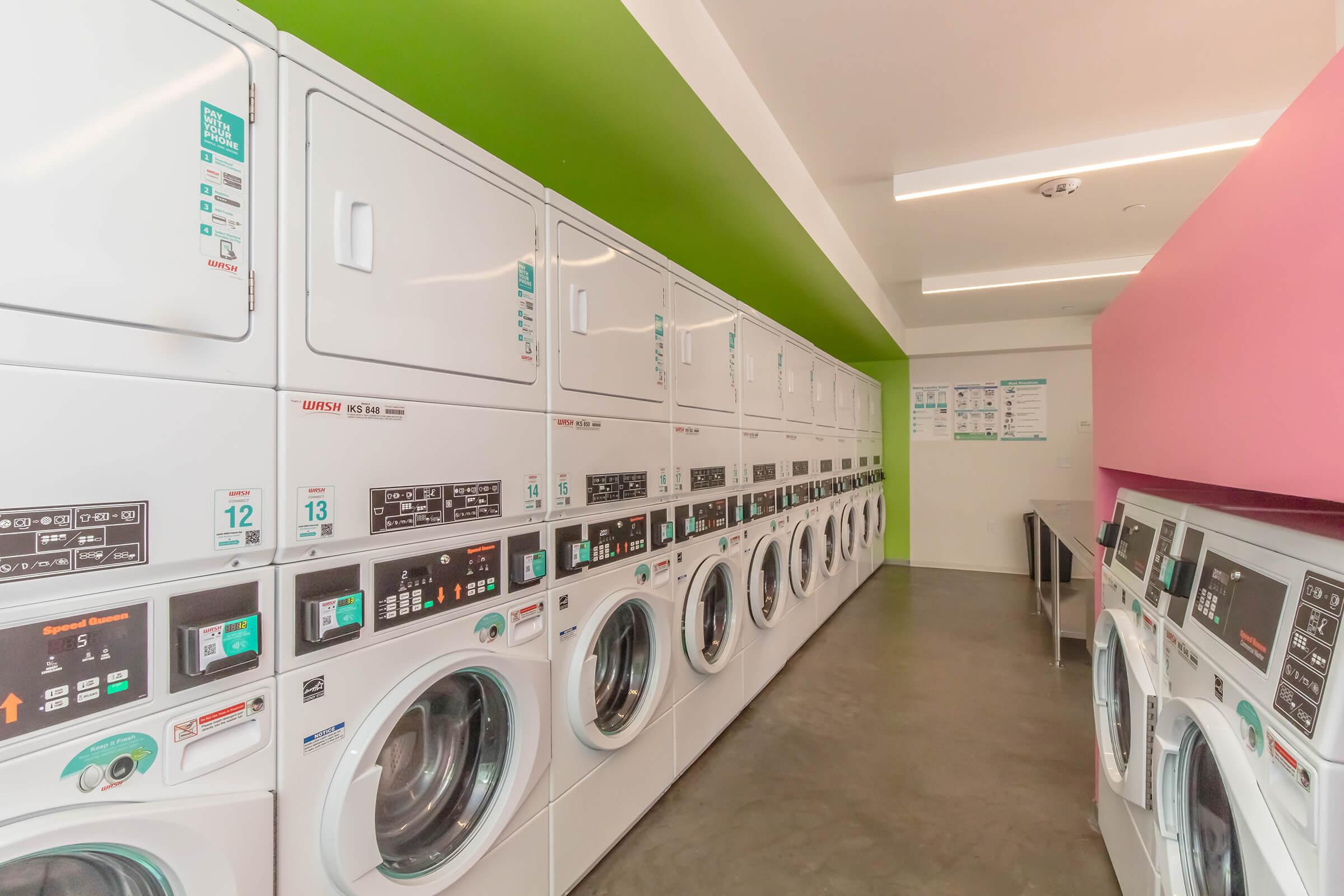 A modern laundromat featuring white washing machines and dryers in a row, with a vibrant green wall on one side and a pink wall on the other. The space has a clean and bright appearance, with various control panels visible on the machines.