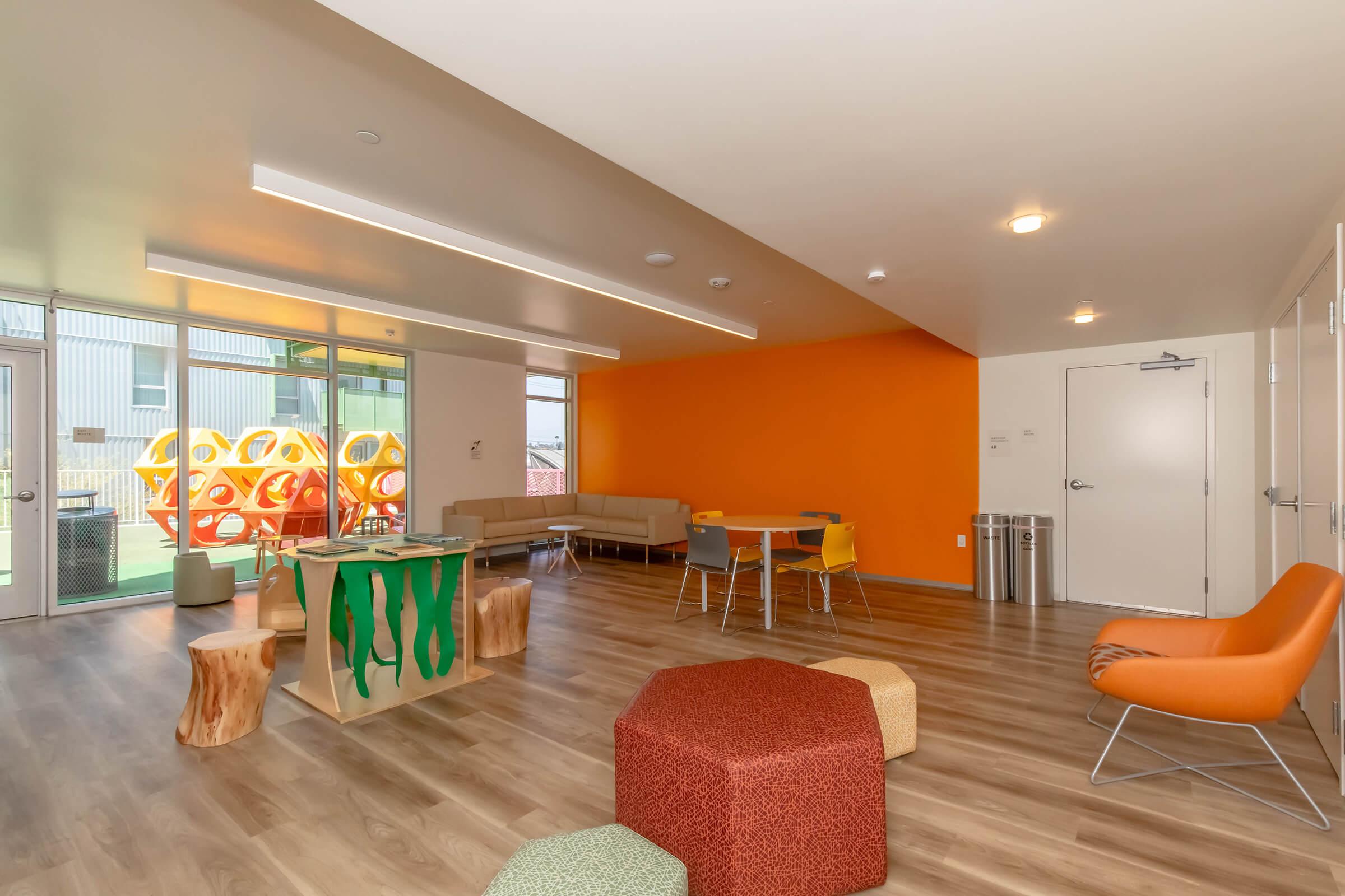 A brightly lit playroom featuring a large orange accent wall, wooden flooring, a modern seating area with colorful ottomans, a table, and a whimsical green and brown play structure. Large windows allow natural light in, enhancing the inviting atmosphere.