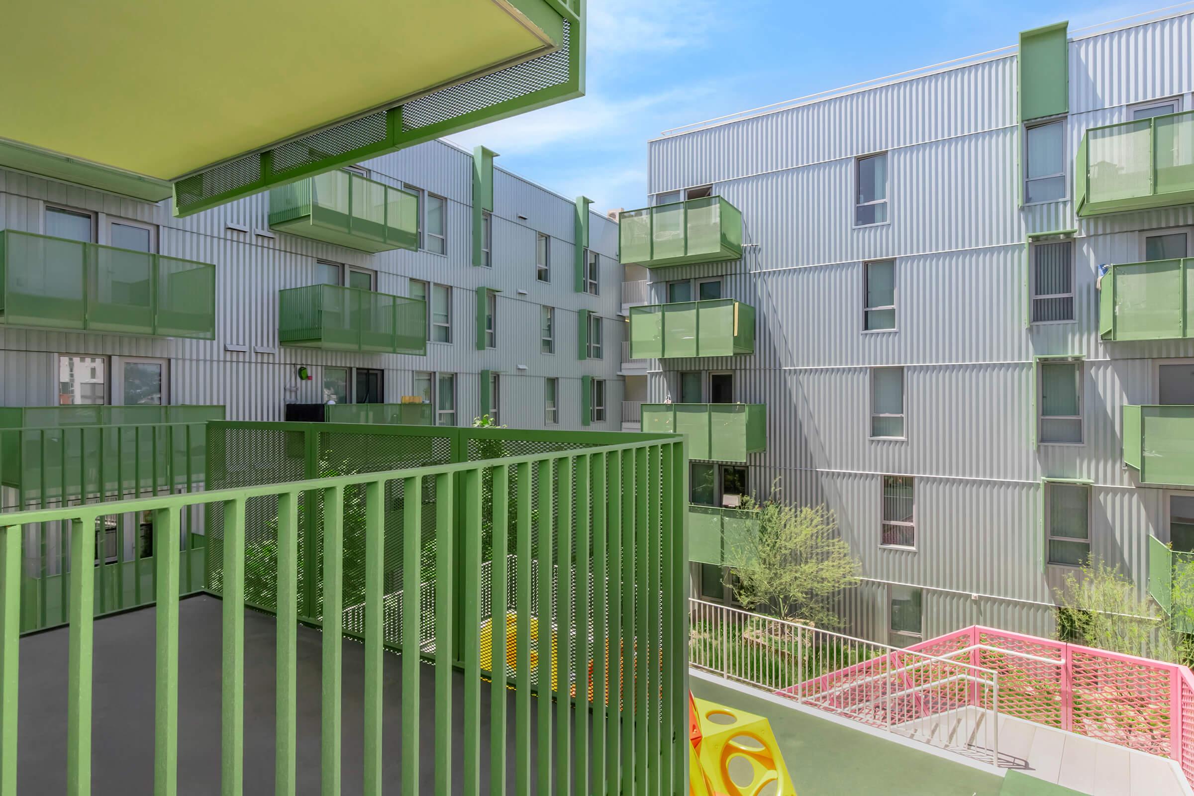 A modern architectural design featuring multiple residential buildings with a sleek, metallic exterior. The buildings have green balconies and railings, surrounded by greenery and colorful play areas. The sky is clear and blue, enhancing the contemporary urban atmosphere.