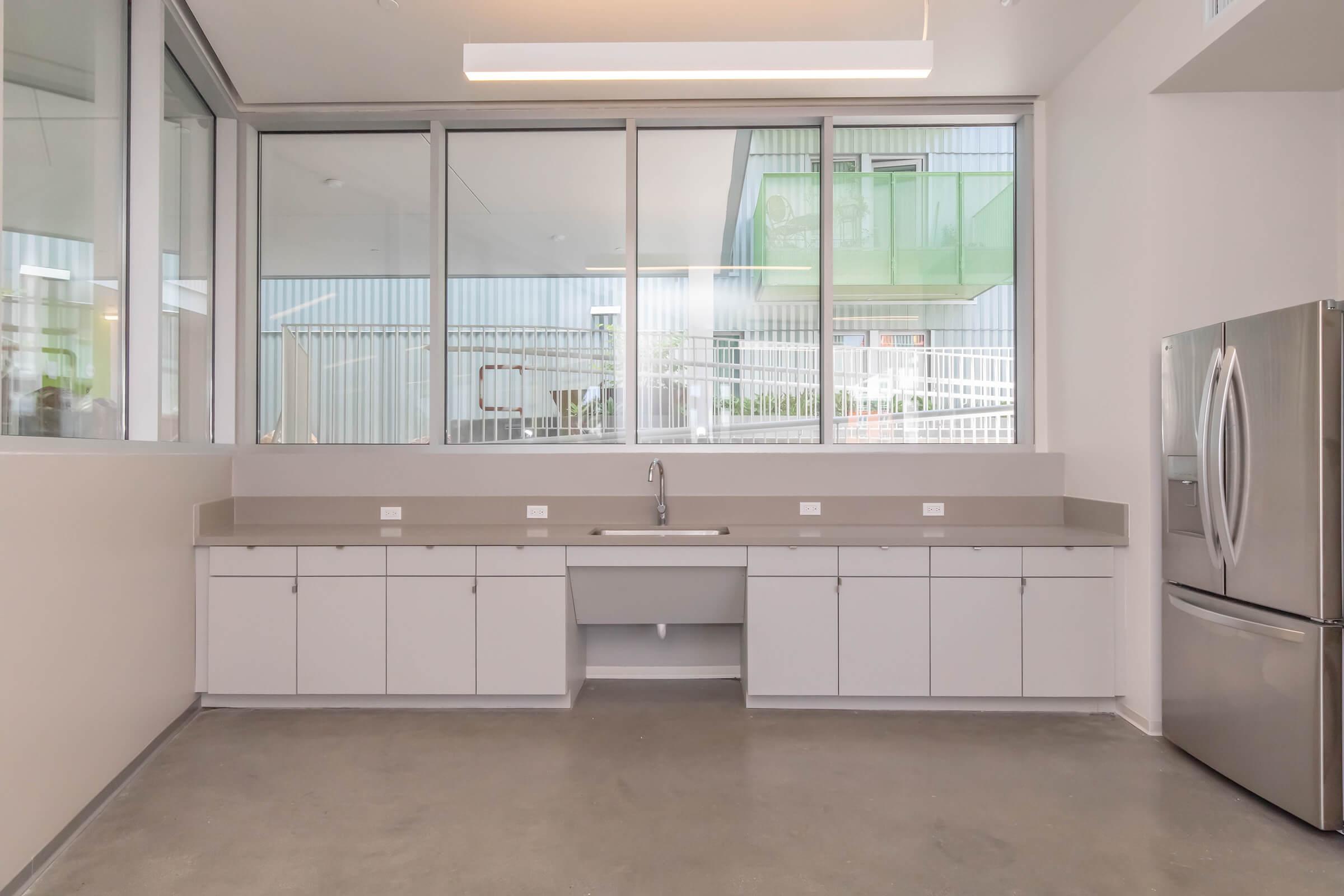 Modern kitchen interior featuring a spacious countertop with a sink, stainless steel refrigerator, and ample white cabinetry. Large windows allow natural light to fill the space, enhancing the contemporary design with a clean, minimalistic aesthetic. The floors are polished concrete, adding to the modern look.