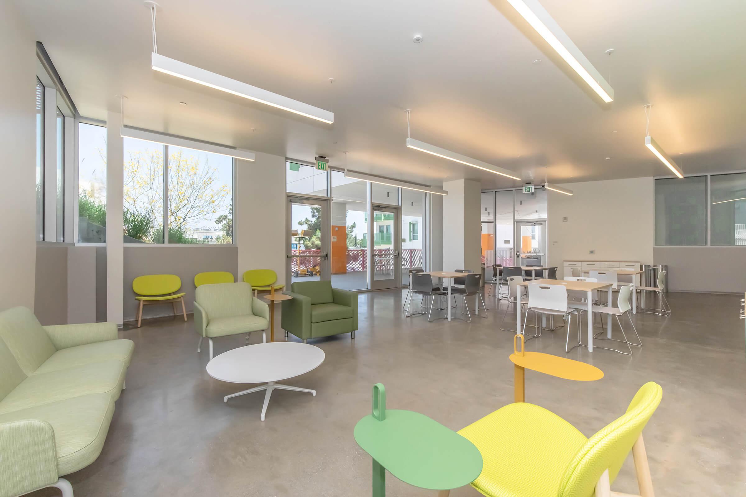 A modern, spacious interior of a communal area featuring various seating options. There are green and yellow chairs, a light-colored sofa, a circular coffee table, and a bright atmosphere created by large windows. Tables and chairs are arranged for group activities, with a view of outdoor greenery.