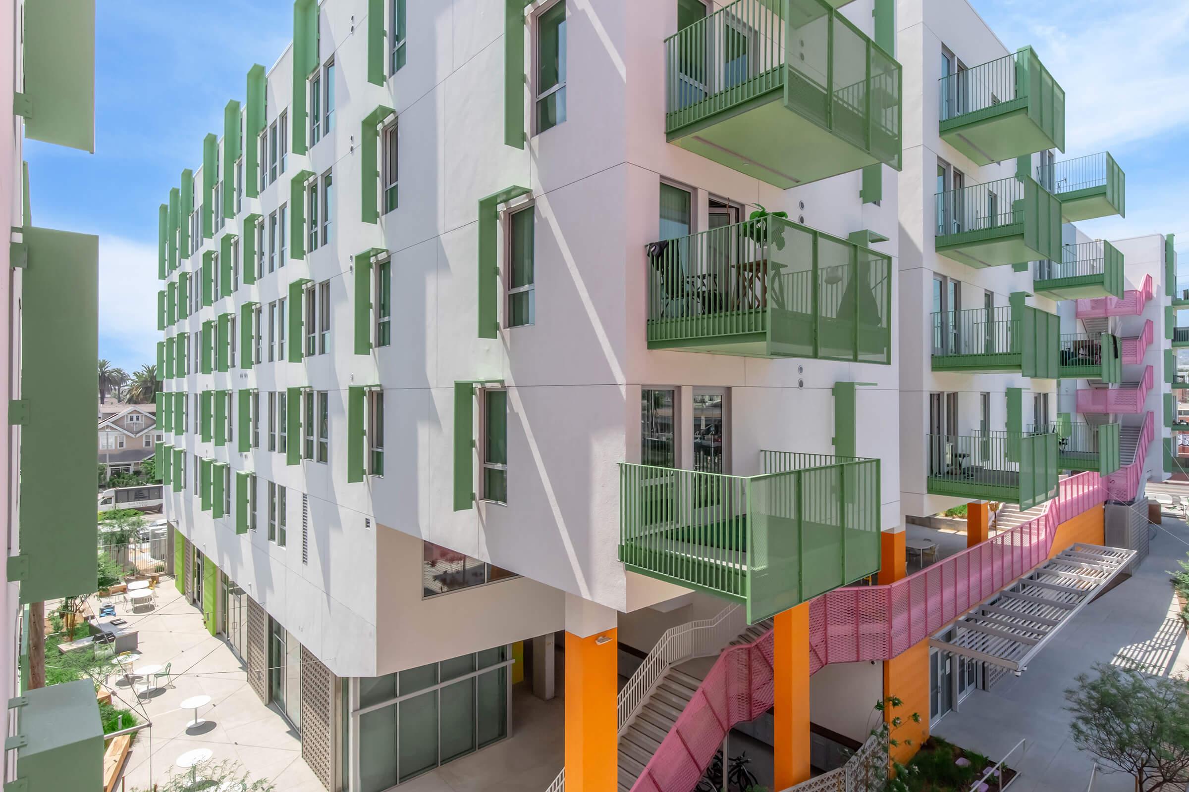 Modern apartment building with a white facade and vibrant green and orange accents. The structure features multiple floors with balconies, and surrounding outdoor areas with landscaping. The design emphasizes contemporary architecture and colorful details, creating an inviting urban living environment.