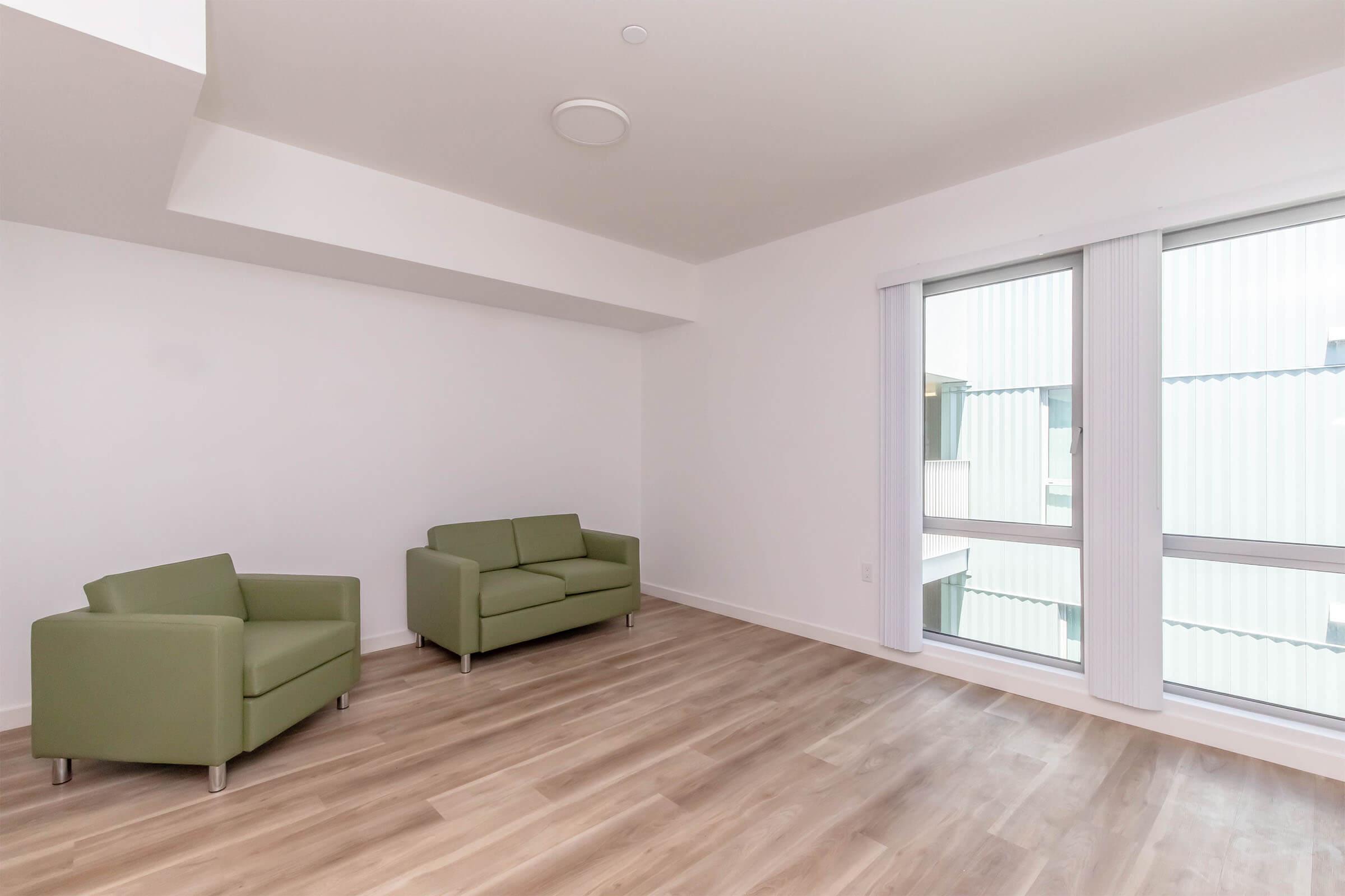 A bright, modern living space featuring two green armchairs and large windows with white blinds. The light wood flooring complements the minimalist design, while the white walls and ceiling create an open, airy atmosphere.