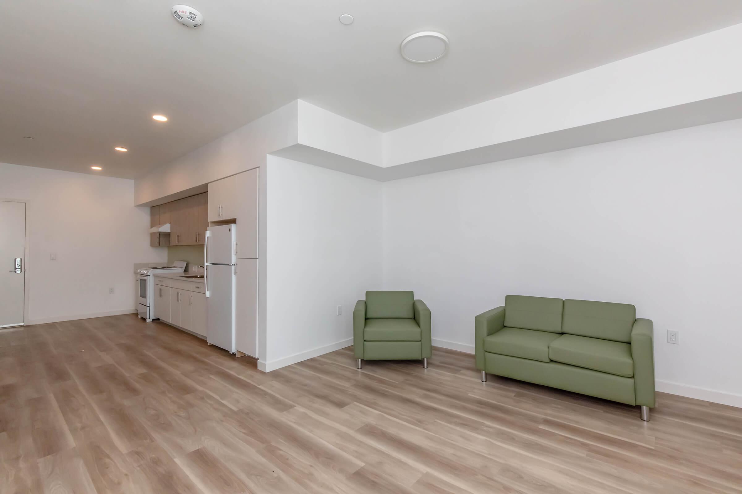A modern, empty room featuring light wood flooring, a small green armchair, and a green sofa. The kitchen area is visible in the background, featuring white cabinets and appliances. The walls are painted white, creating a bright and spacious atmosphere.