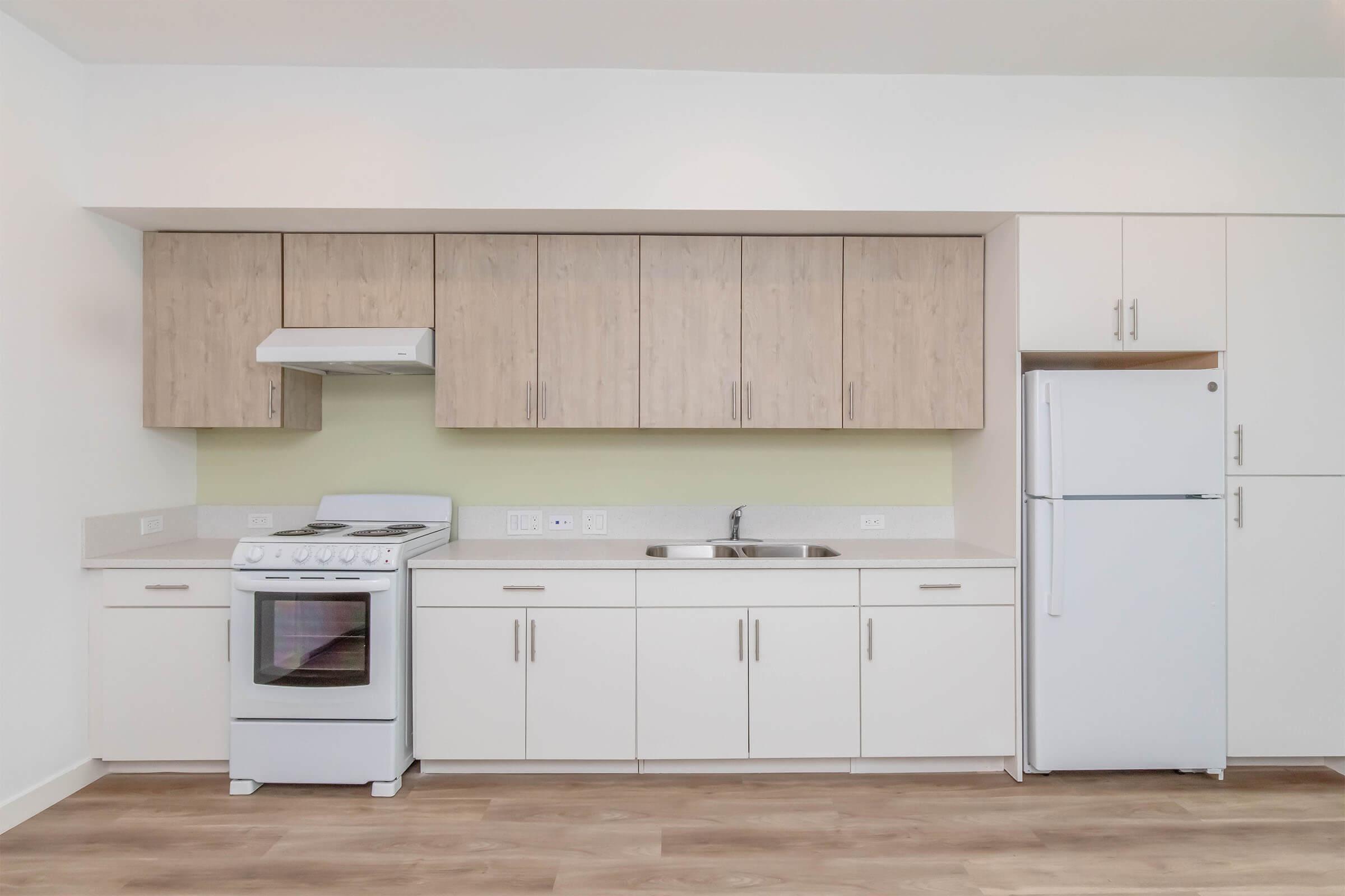 A modern kitchen featuring light wood cabinetry, a white refrigerator, and a white stove. The countertops are light-colored, and there's a stainless steel sink. The walls are painted a soft pale color, creating a bright and airy atmosphere. Natural light illuminates the space through an unseen window.
