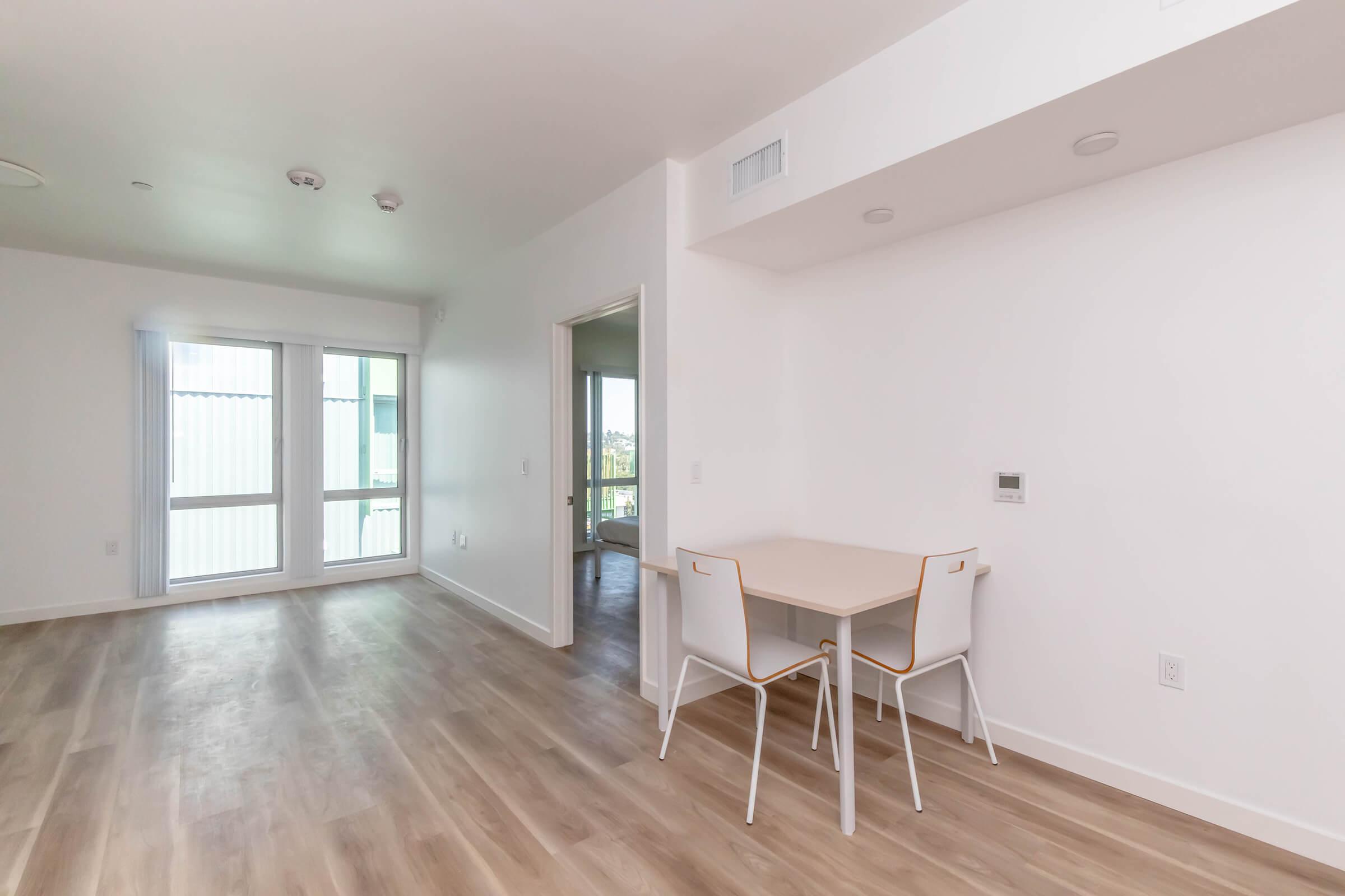 Light and airy modern apartment interior featuring a small dining table with two chairs, large windows allowing natural light, and light wood flooring. The walls are painted white, and there are minimalist design elements throughout the space.