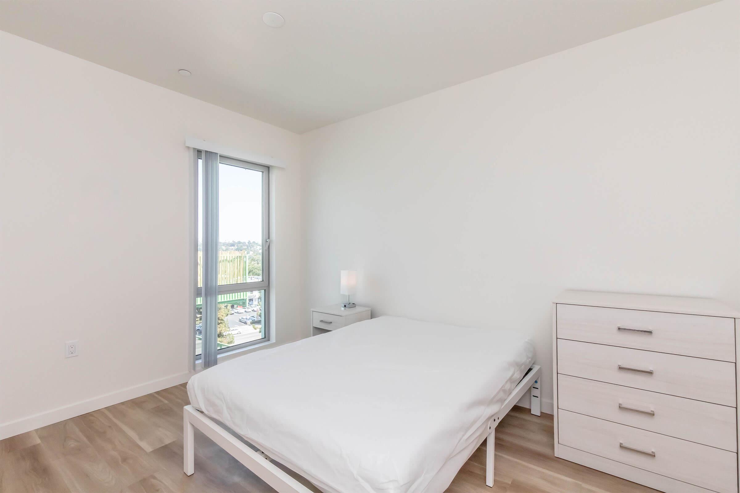 A minimalist bedroom featuring a white bed with a plain white duvet, a bedside lamp on a small nightstand, and a light-colored dresser. Large windows let in natural light, revealing a view outside. The walls and flooring are neutral tones, contributing to a clean and simple aesthetic.