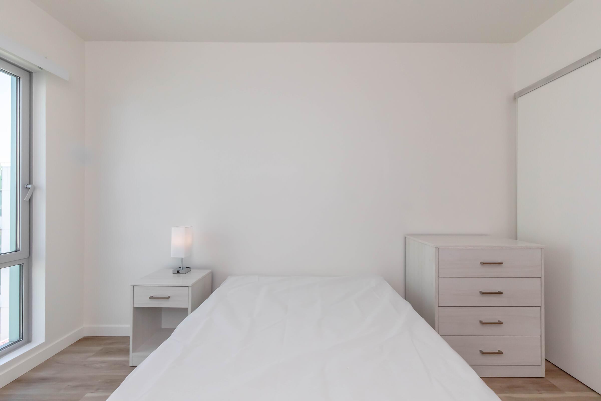 A minimalist bedroom featuring a white bed with an unmade mattress, a bedside table with a lamp, and a white dresser. The room has neutral-colored walls and a large window allowing natural light to enter, creating a bright and airy atmosphere.