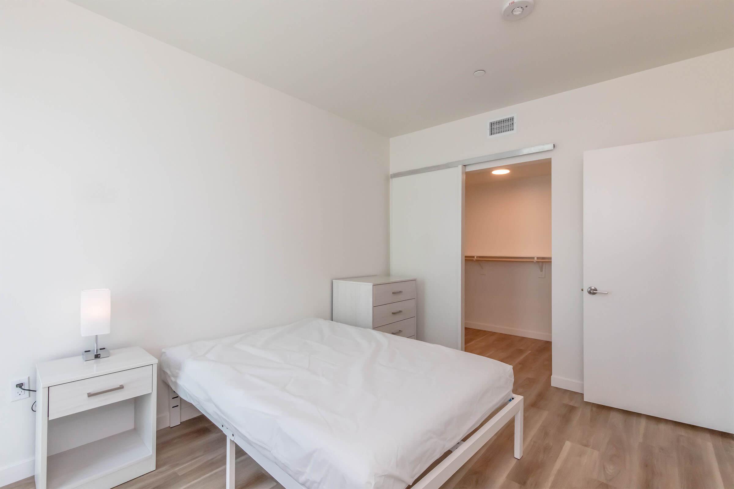 A minimalistic bedroom featuring a white bed frame with a mattress, a small bedside table with a lamp, a dresser, and a closet with sliding doors. The walls are painted white, and the flooring is a light wood. A door leads to a space that appears to be a closet or storage area.